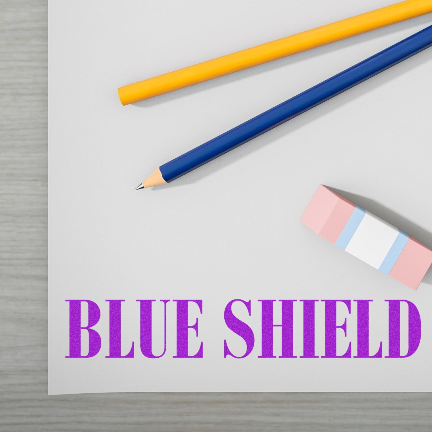 Blue Shield Rubber Stamp on paper with two pencils, one yellow and one blue, and a pink eraser with blue and white stripes.