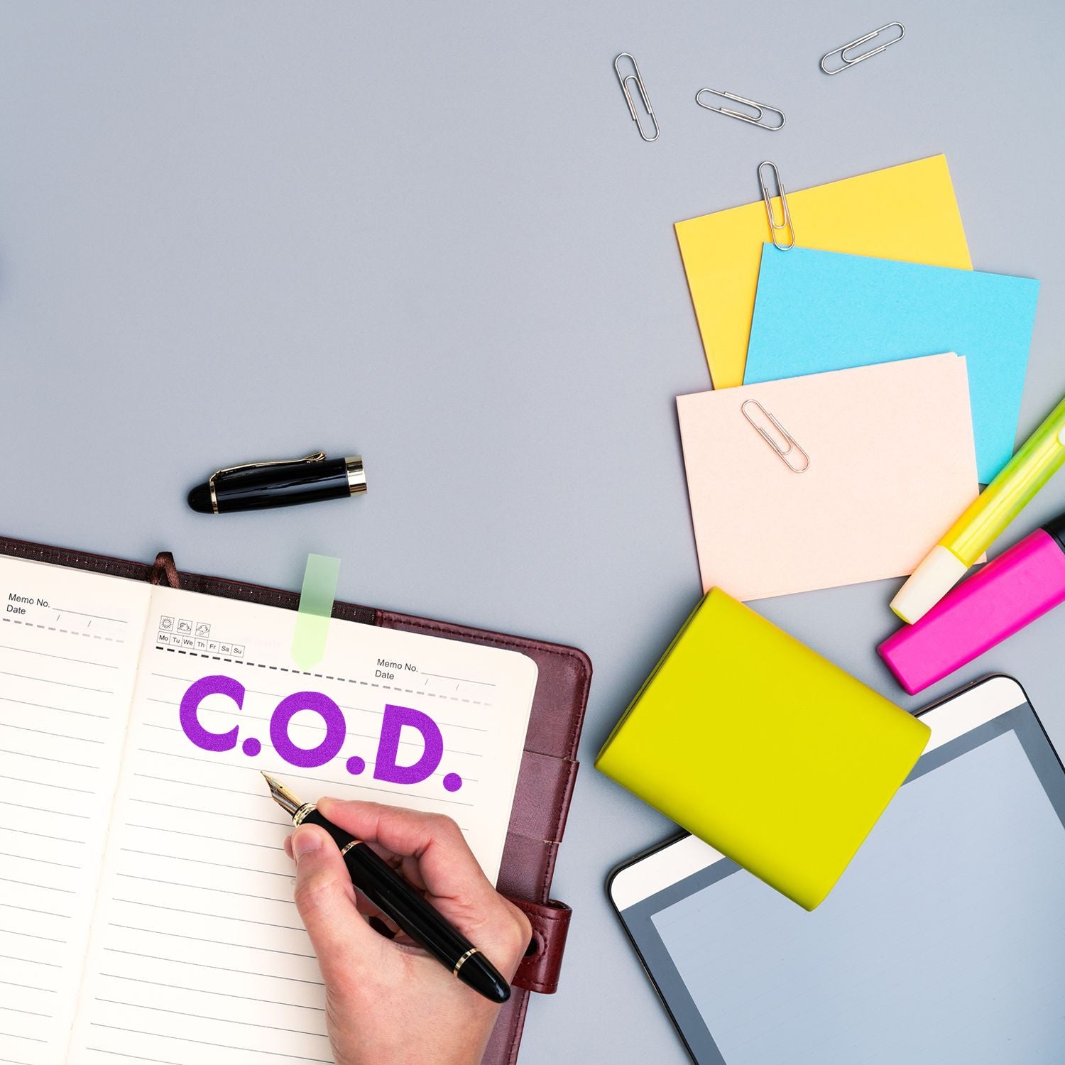 Hand using Large Self Inking Bold C.O.D. Stamp on a notebook, surrounded by colorful sticky notes, paper clips, a pen, and a tablet.