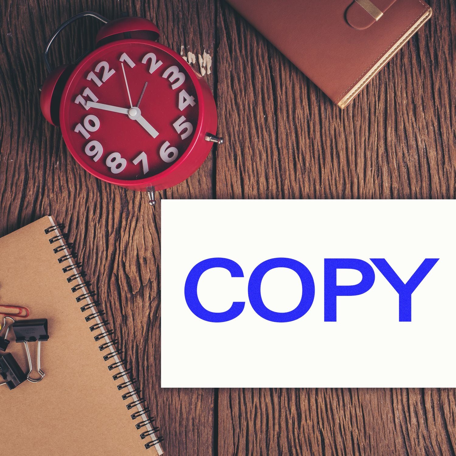 A red clock, brown notebook, and paper stamped with COPY in blue ink using the Self Inking Bold Copy Stamp on a wooden desk.