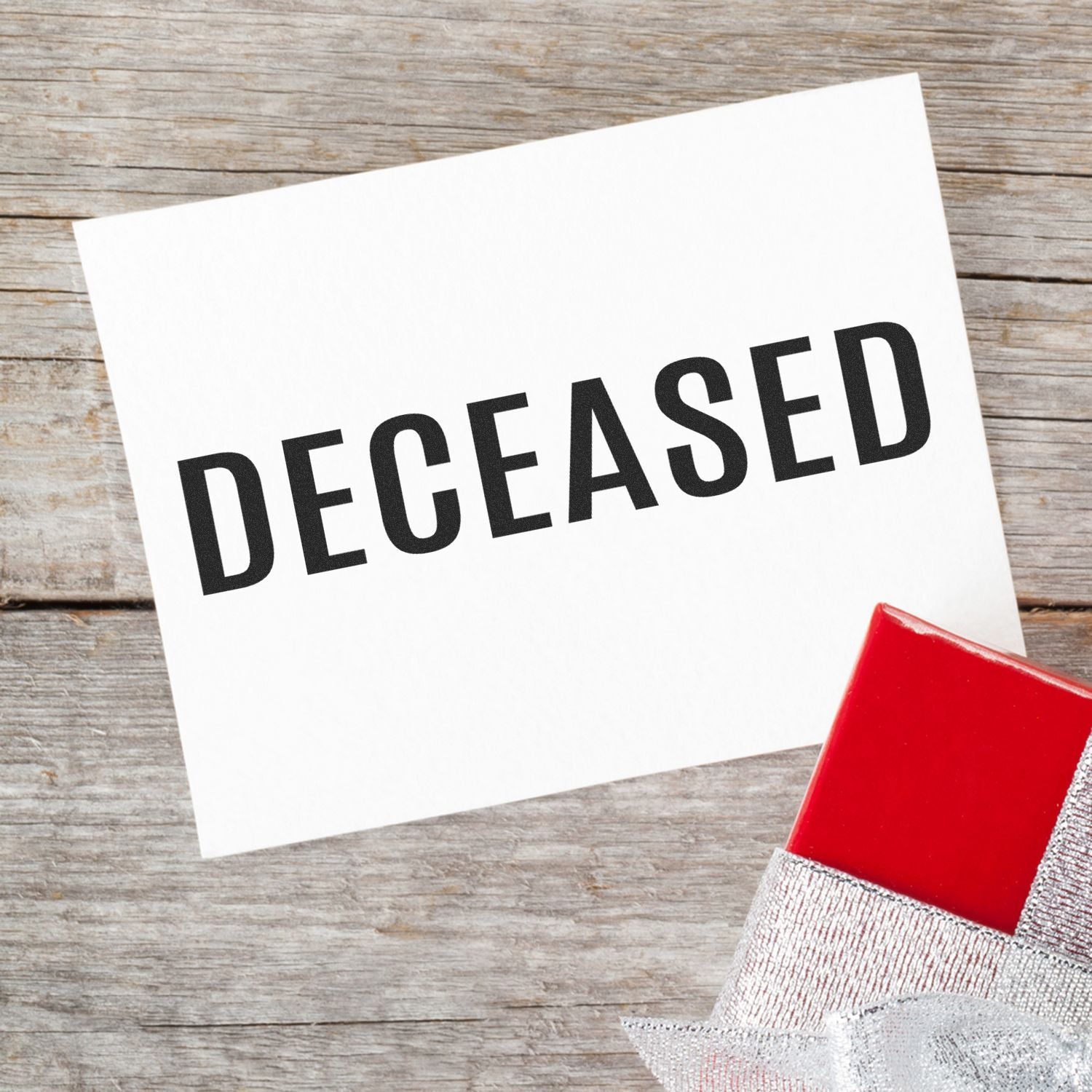 White card stamped with DECEASED using the Large Pre-Inked Bold Deceased Stamp, placed on a wooden surface next to a red gift box.