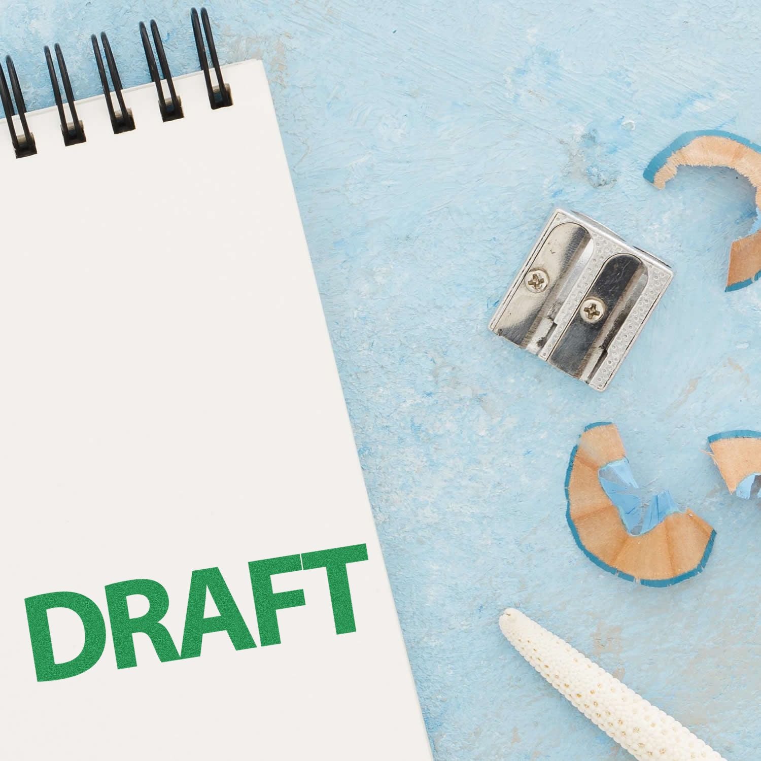 Notepad with DRAFT in green text, pencil sharpener, and shavings on a light blue surface. Focus word: Bold Draft Rubber Stamp.