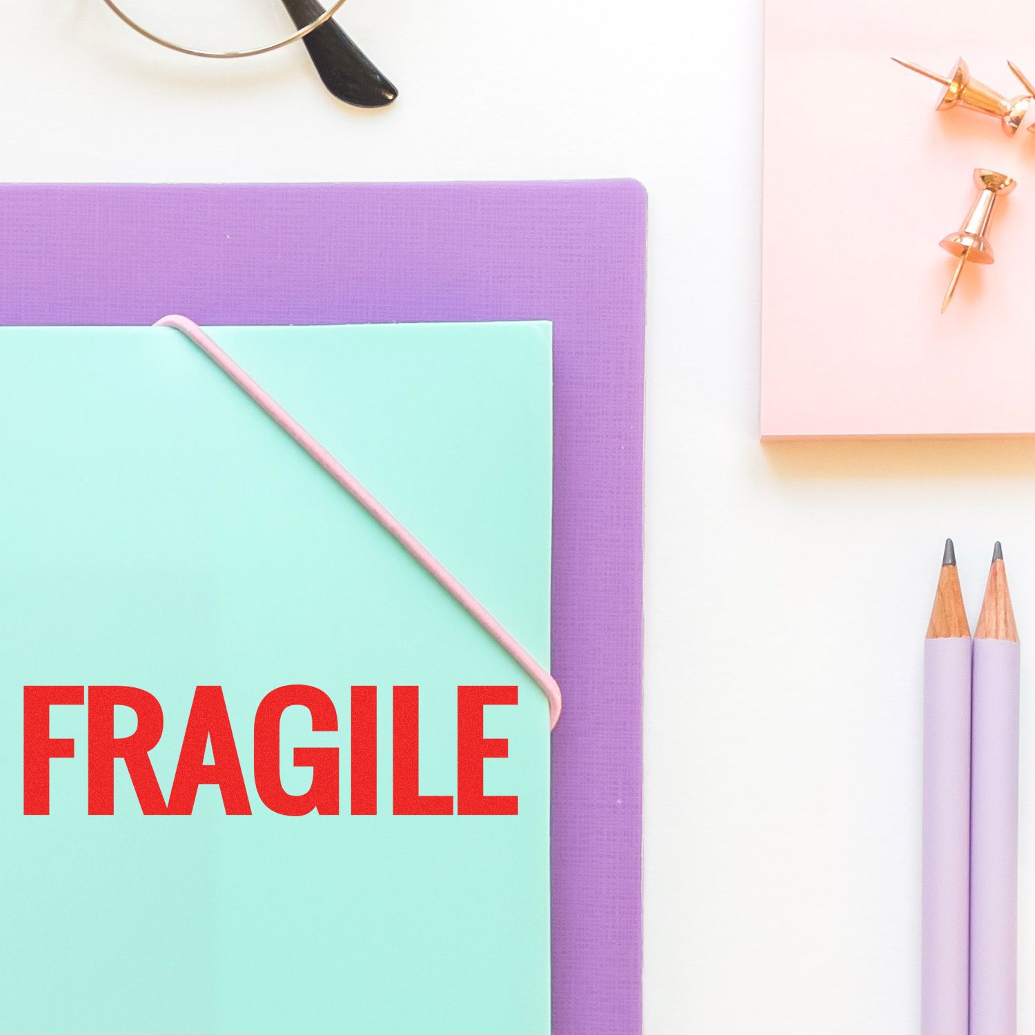 Self Inking Bold Fragile Stamp used on a mint green envelope, placed on a purple folder, with pencils and push pins nearby.