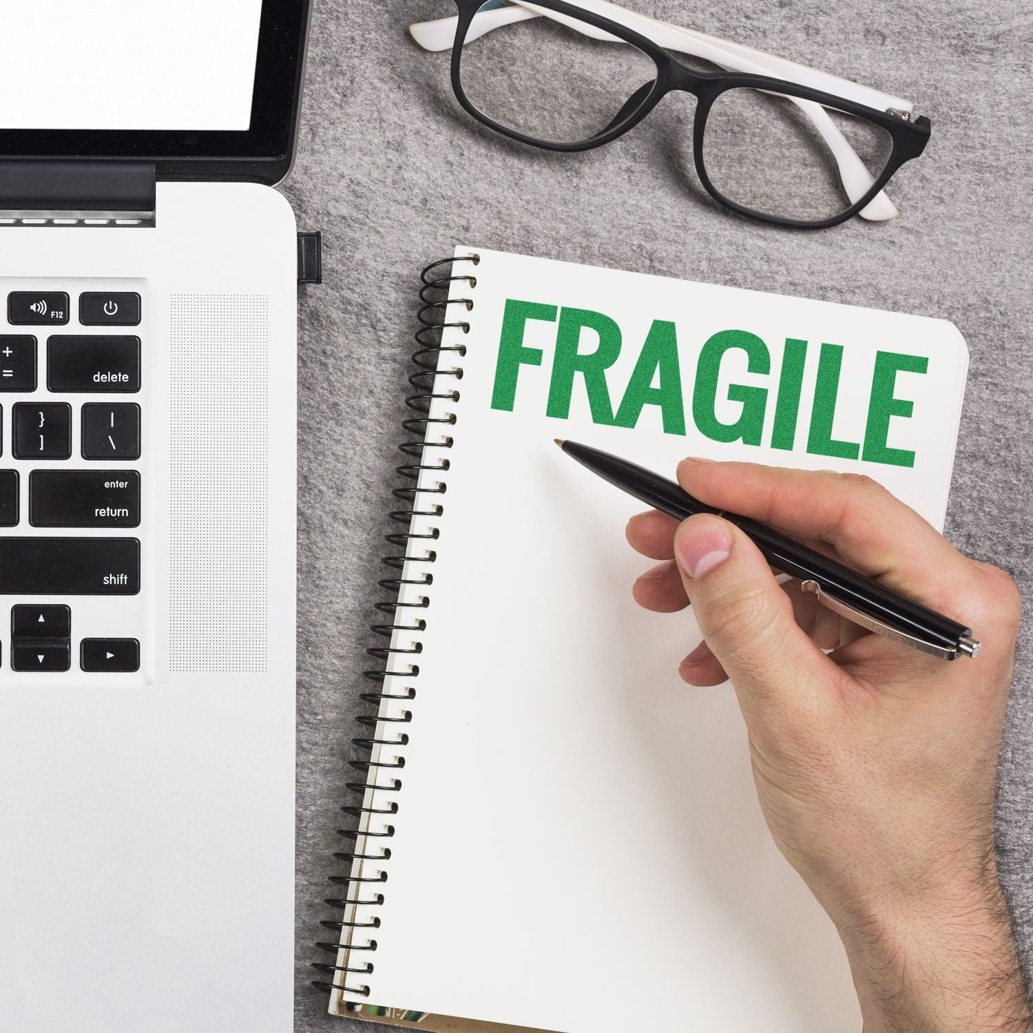 A hand holding a pen near a notebook stamped with FRAGILE using the Bold Fragile Rubber Stamp, next to a laptop and glasses.