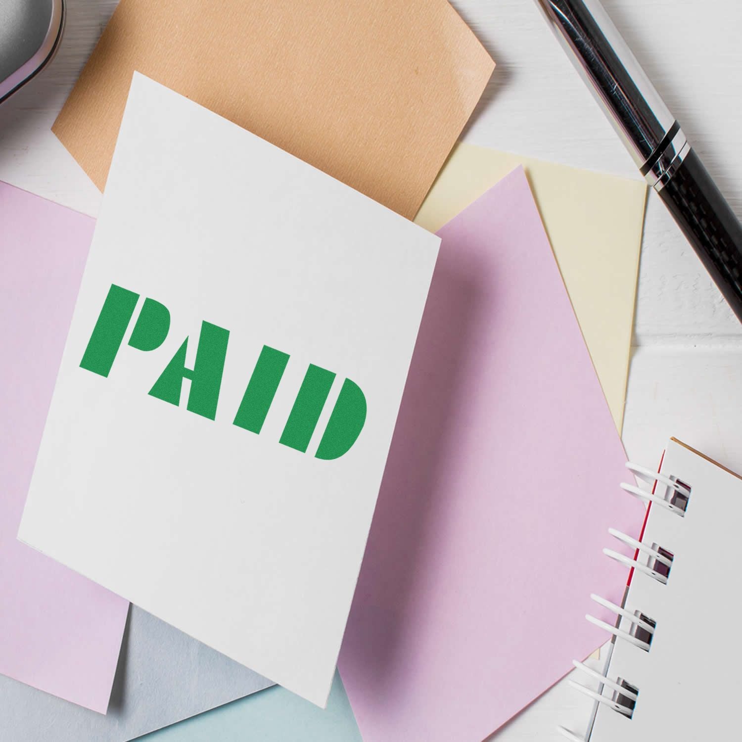 Bold Paid Rubber Stamp on a white card surrounded by pastel-colored papers, a pen, and a notebook.