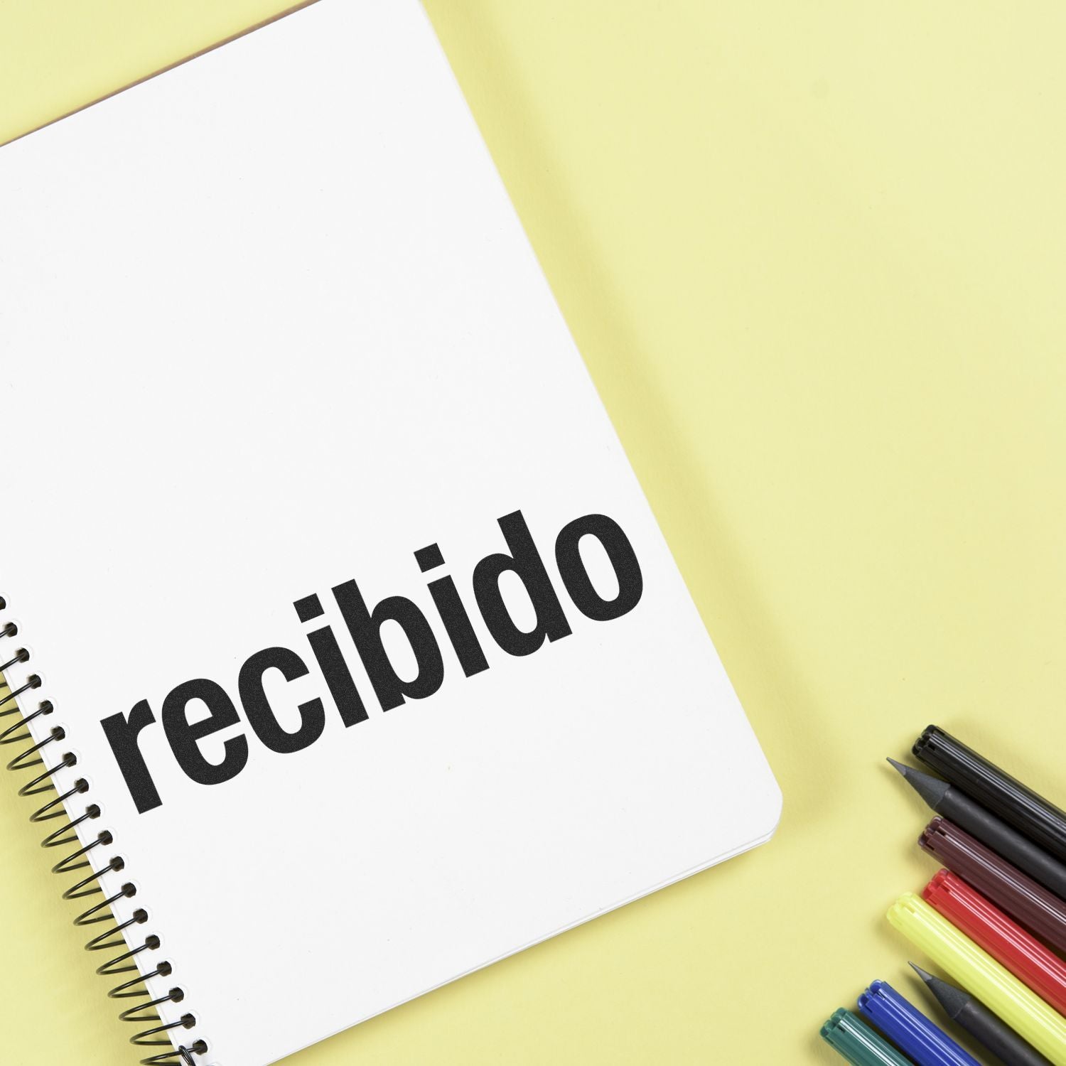 Large Bold Recibido Rubber Stamp used on a white notebook, with colorful pens placed beside it on a yellow background.