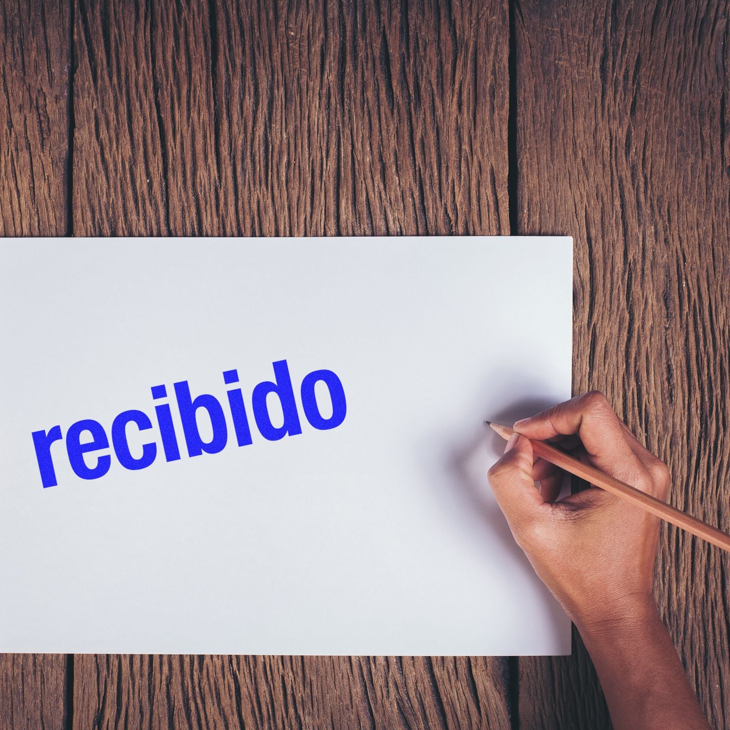 Hand holding a pencil next to a paper stamped with recibido in blue ink using the Bold Recibido Rubber Stamp on a wooden surface.