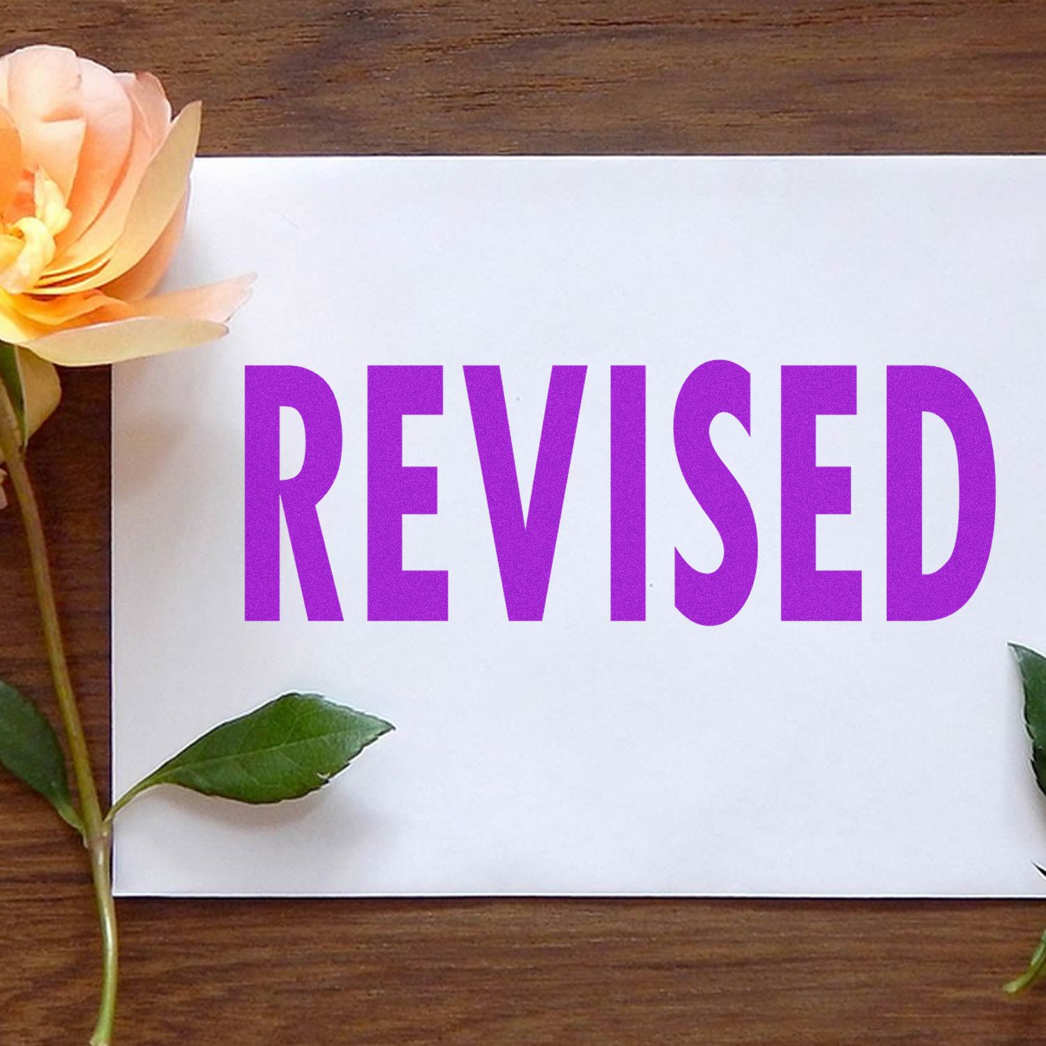 Self Inking Bold Revised Stamp in use, displaying the word REVISED in bold purple letters on a white paper with a flower beside it.