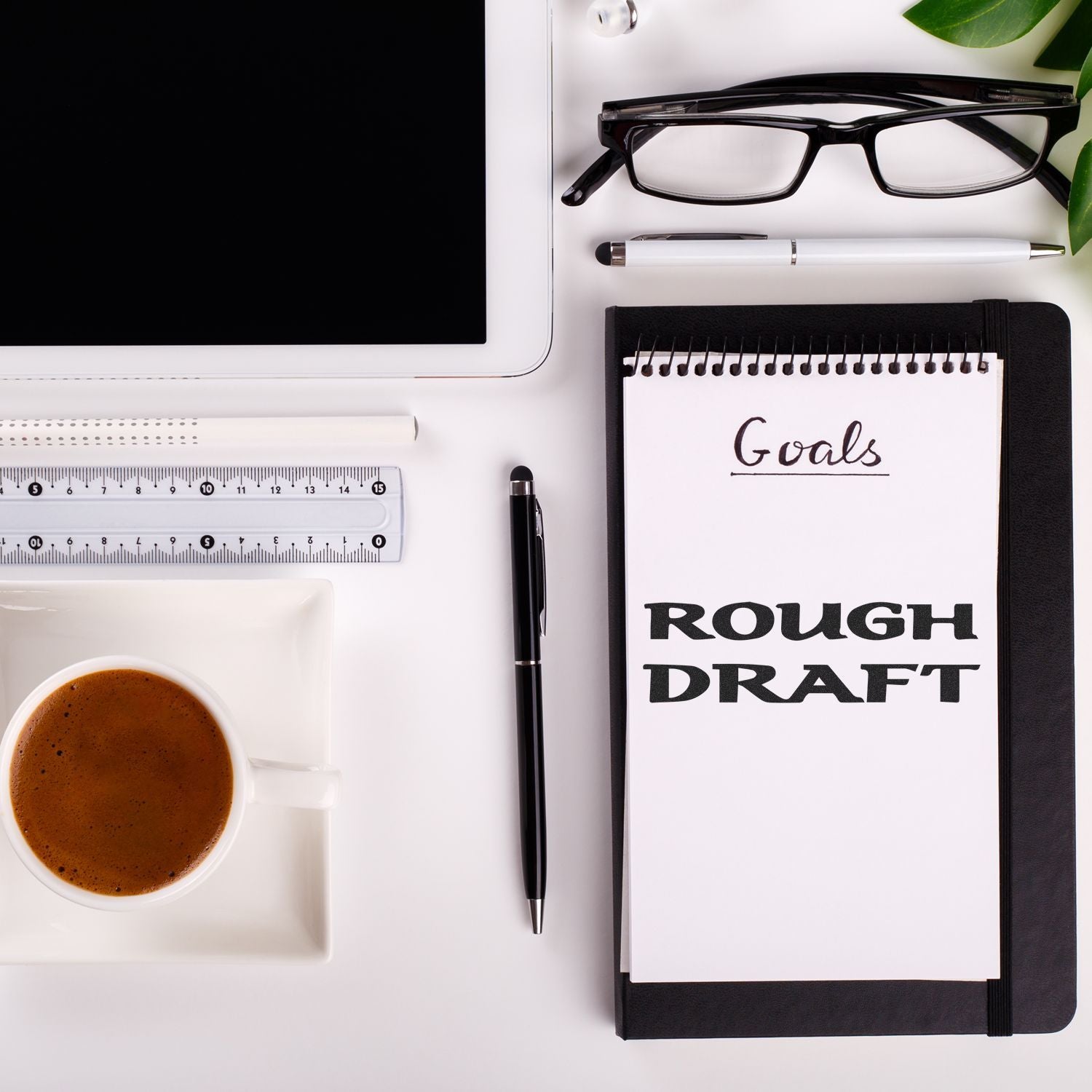 Desk setup with a Large Bold Rough Draft Rubber Stamp imprint on a notepad, surrounded by a tablet, glasses, ruler, pen, and coffee.