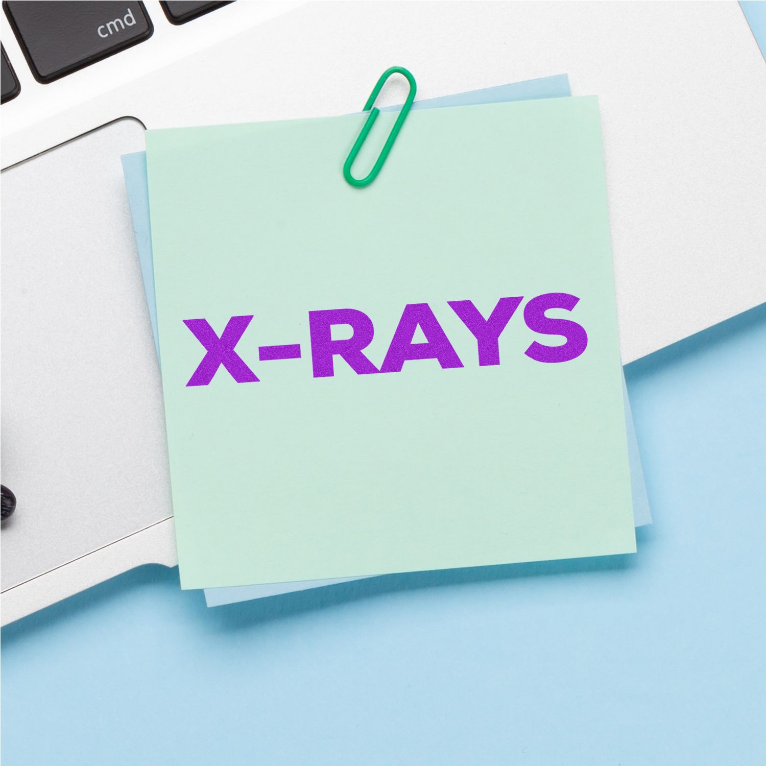 A mint green sticky note stamped with X-RAYS in bold purple ink from the Large Self Inking Bold X-Rays Stamp, clipped to a laptop.