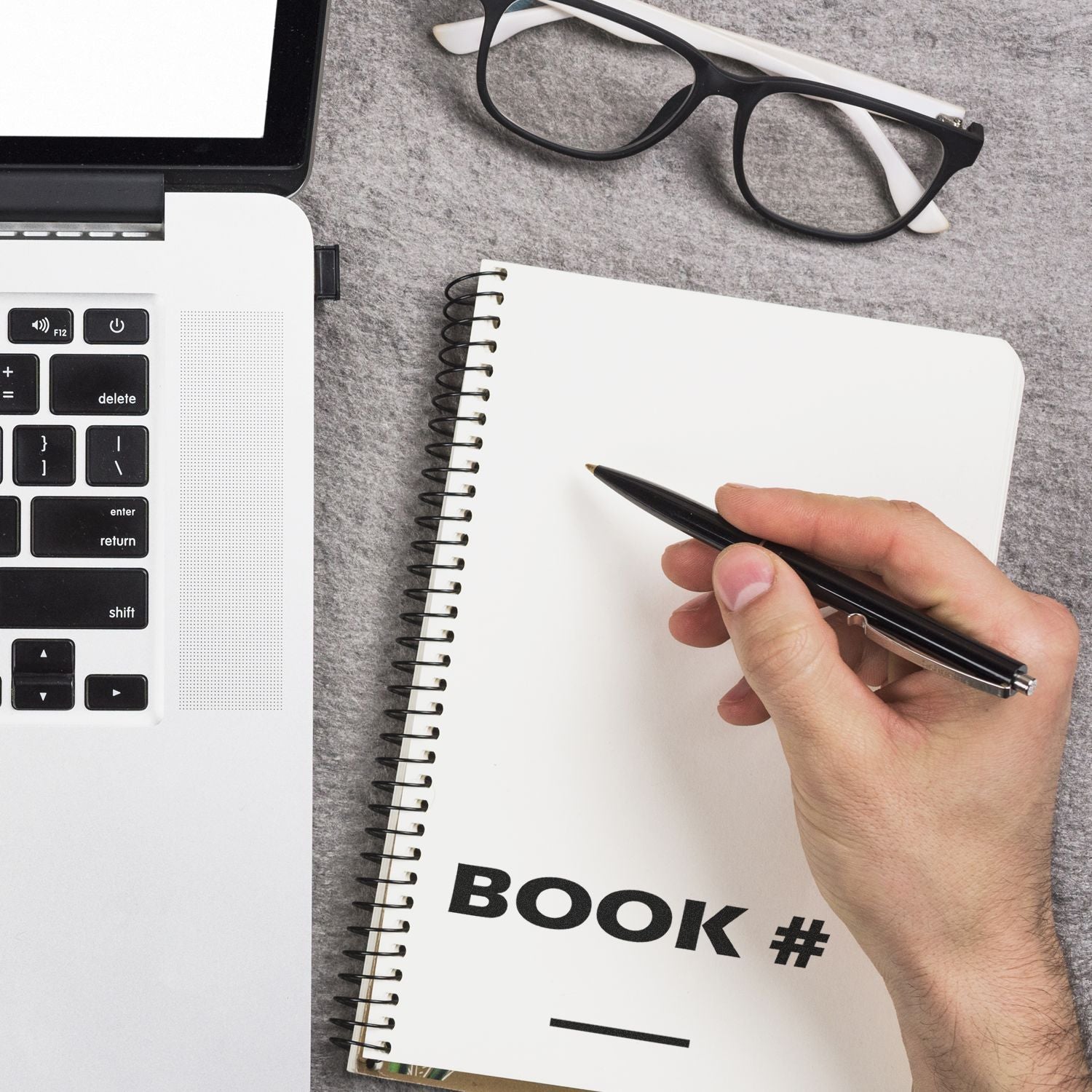 Hand using the Large Book Rubber Stamp on a spiral notebook near a laptop and glasses, with a pen ready to write.