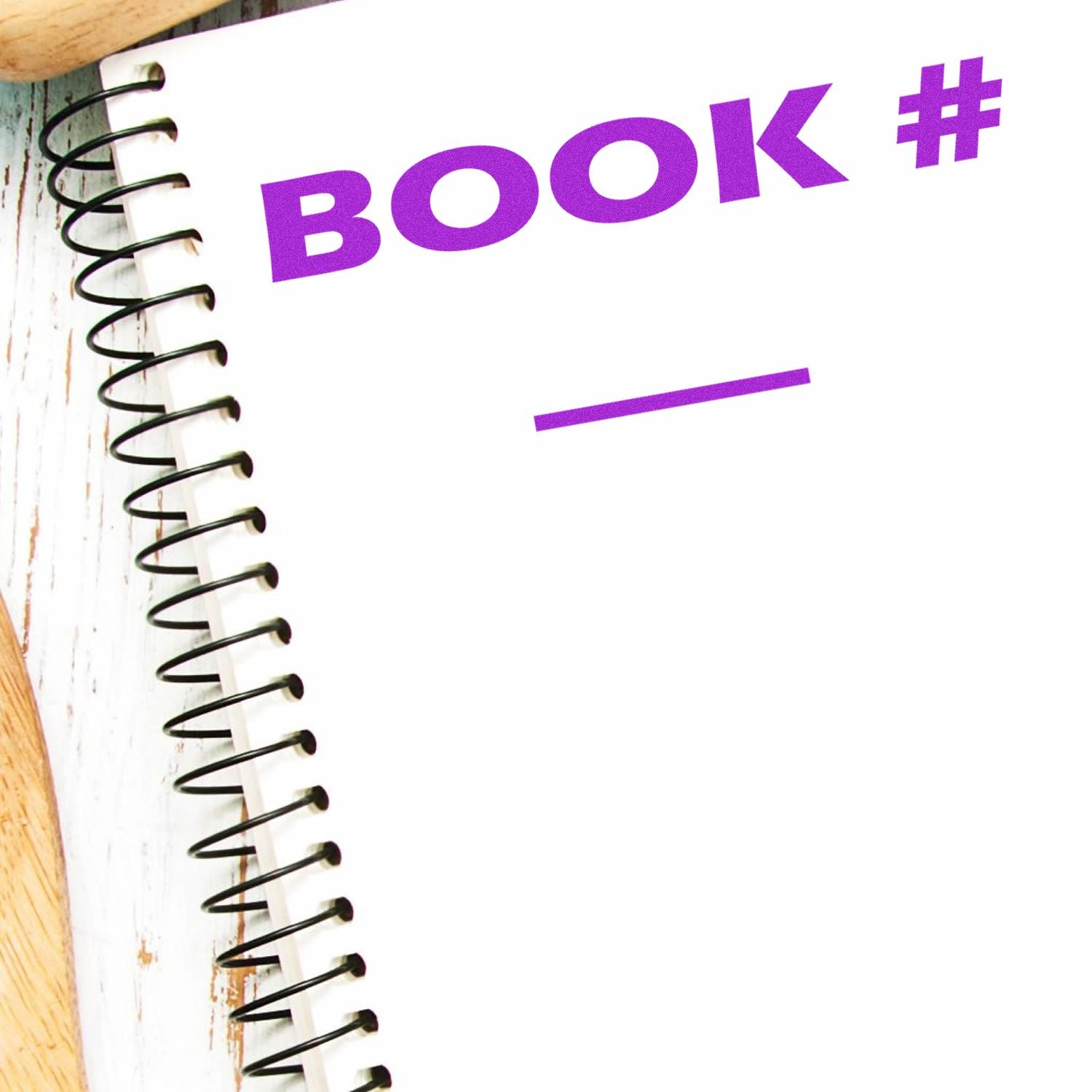 Spiral notebook with a purple BOOK # imprint from the Book Rubber Stamp on a white page, placed on a wooden surface.