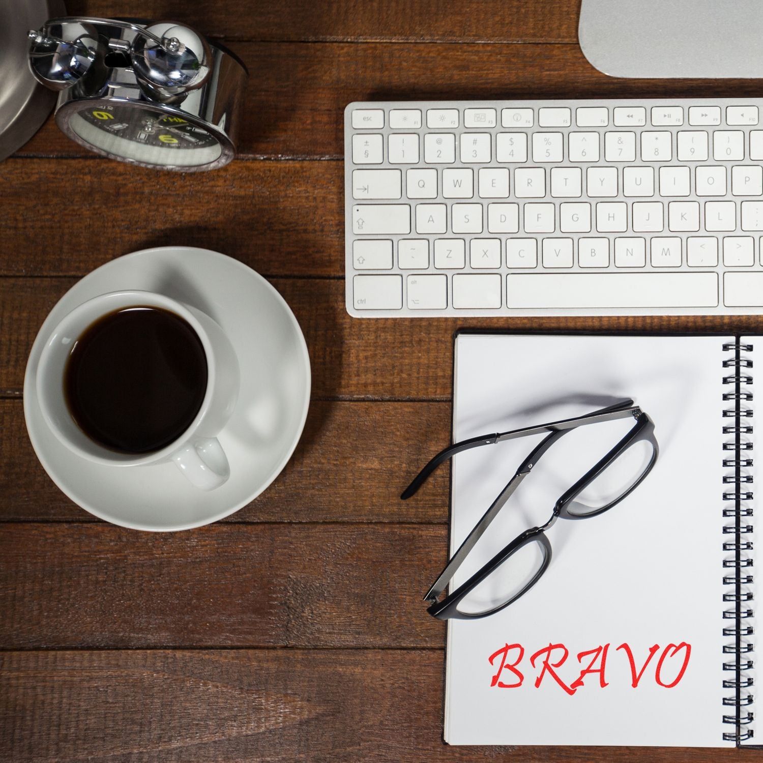 A desk with a keyboard, coffee cup, glasses, notebook, and Large Bravo Rubber Stamp imprinting BRAVO in red on the notebook.