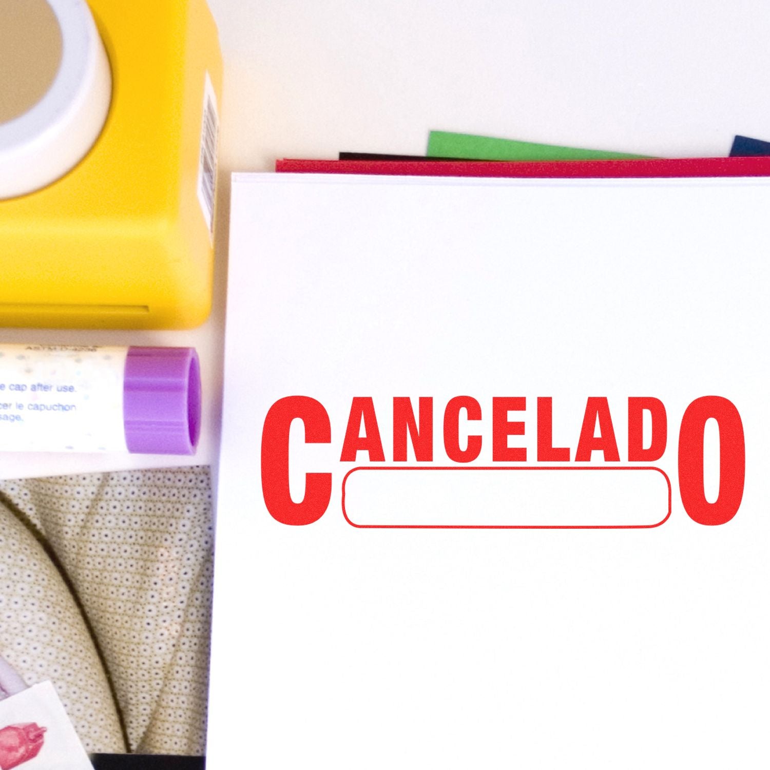 Cancelado with Box Rubber Stamp in red ink on white paper, surrounded by office supplies including a yellow box and a purple glue stick.