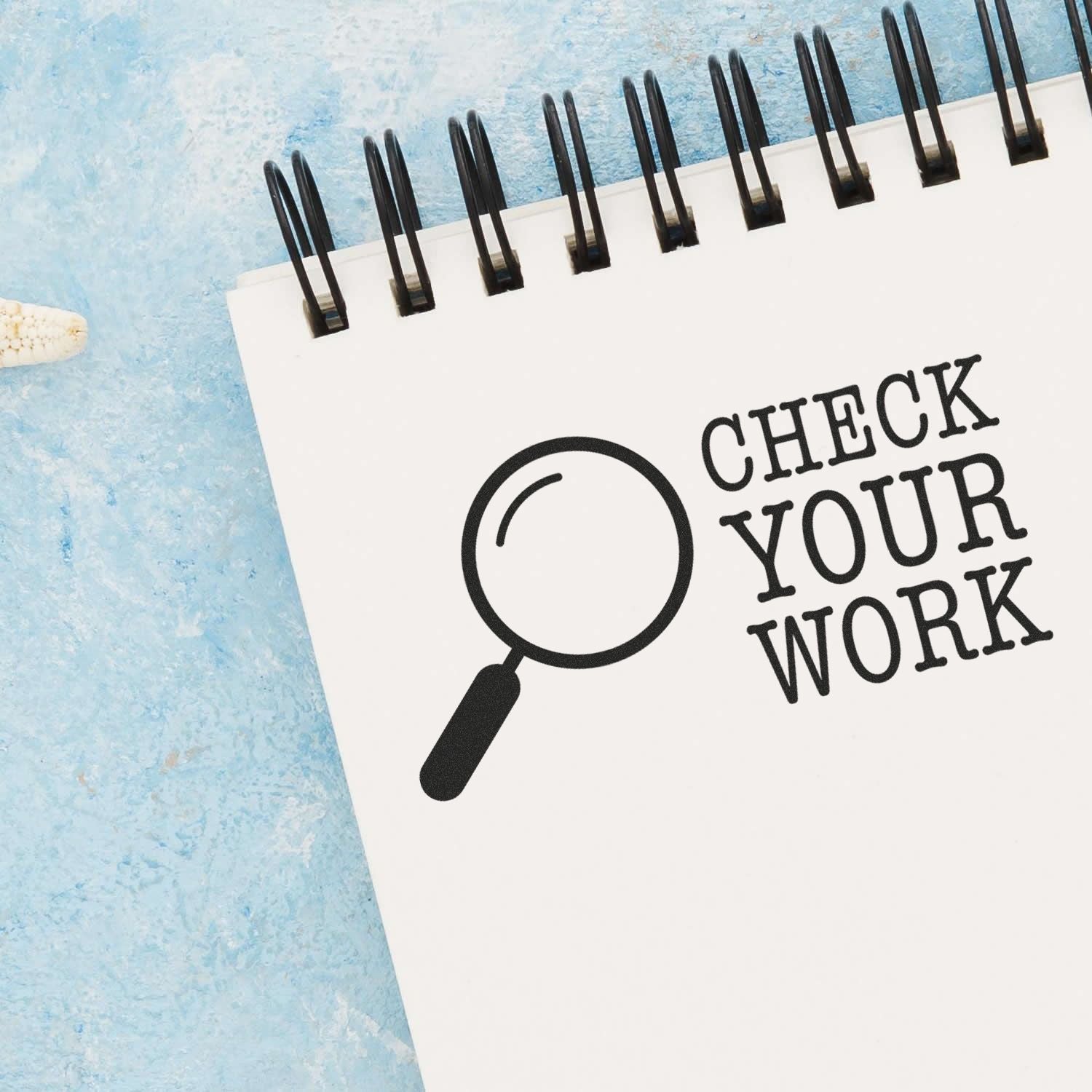 A Check Your Work rubber stamp imprint on a spiral notebook page with a magnifying glass icon, placed on a light blue textured surface.