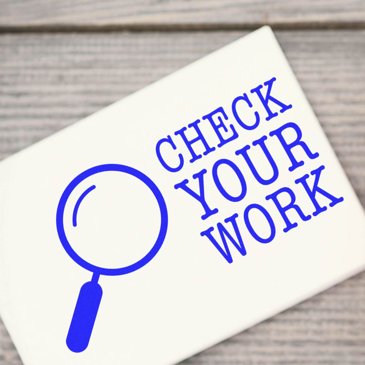 Large Check Your Work Rubber Stamp in use, featuring a magnifying glass icon and bold blue text on a white background, placed on a wooden surface.