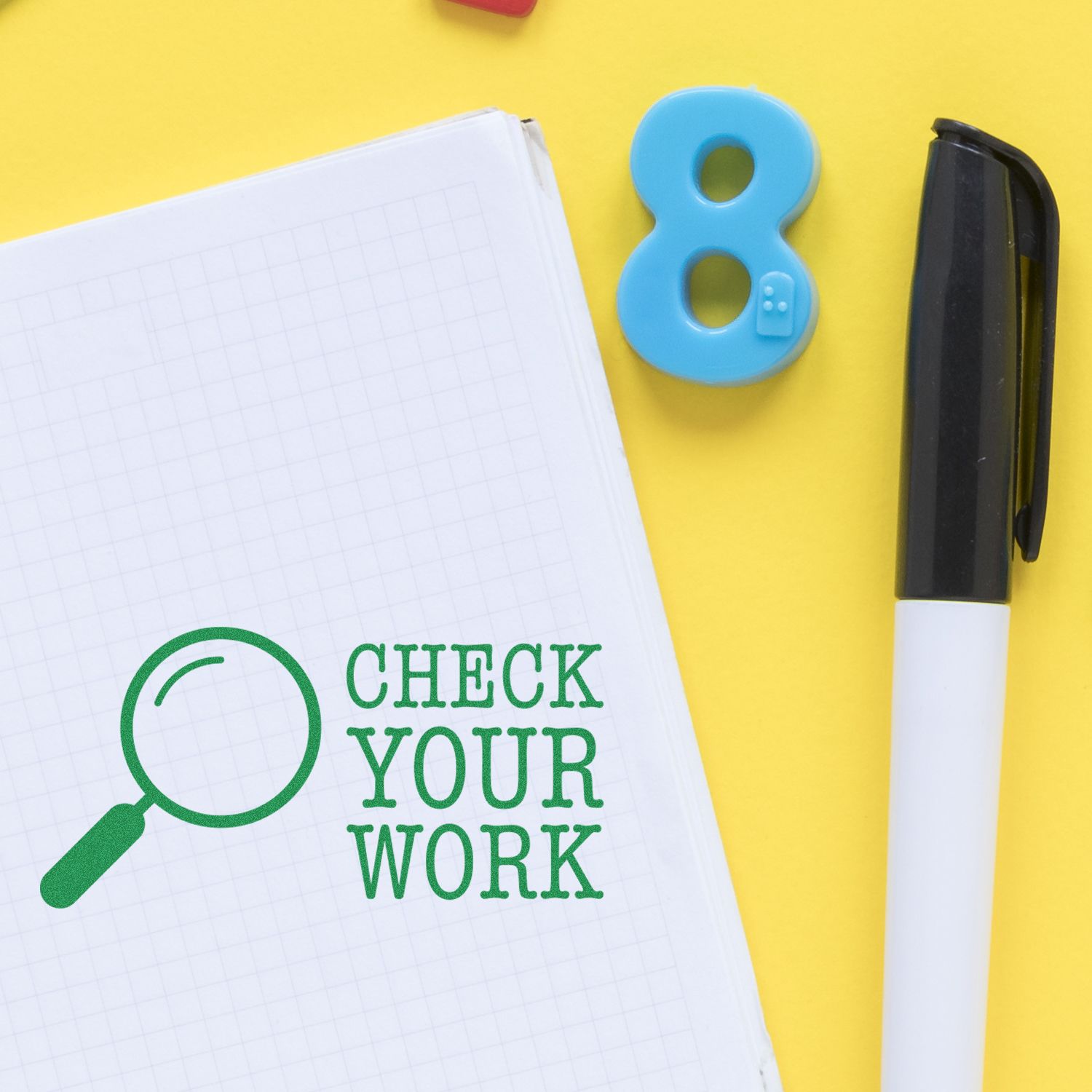 Large Check Your Work Rubber Stamp in green ink on graph paper, next to a black pen and a blue number 8 on a yellow background.