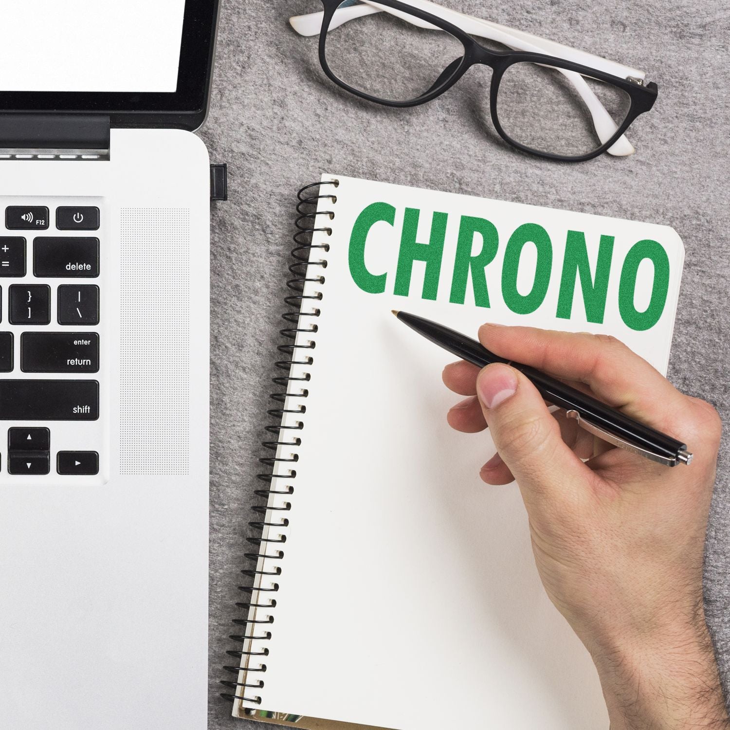 Hand writing in a notebook with CHRONO stamped on it using the Chrono Rubber Stamp, next to a laptop and glasses on a gray surface.