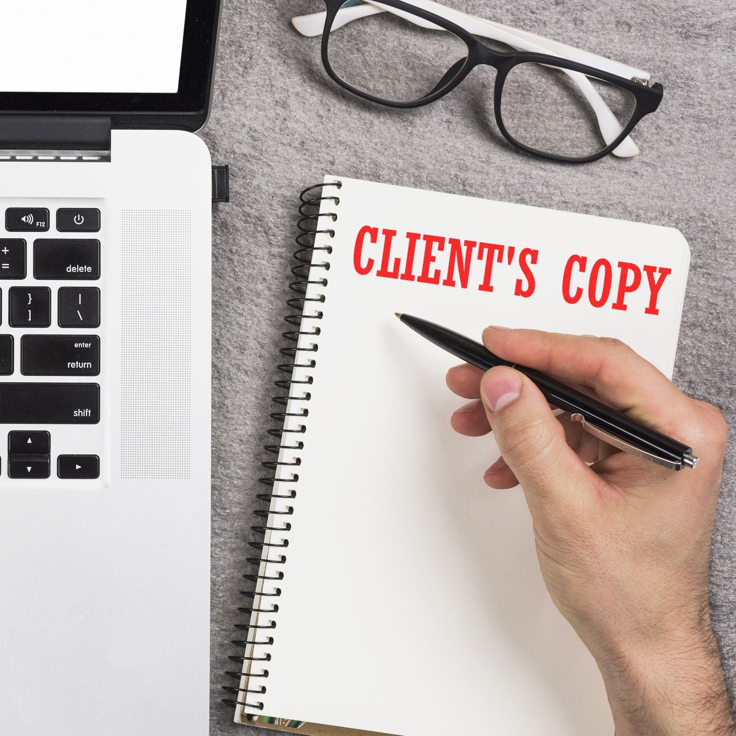 Hand writing in a notebook labeled CLIENT'S COPY next to a laptop, glasses, and a Clients Copy Rubber Stamp on a gray surface.