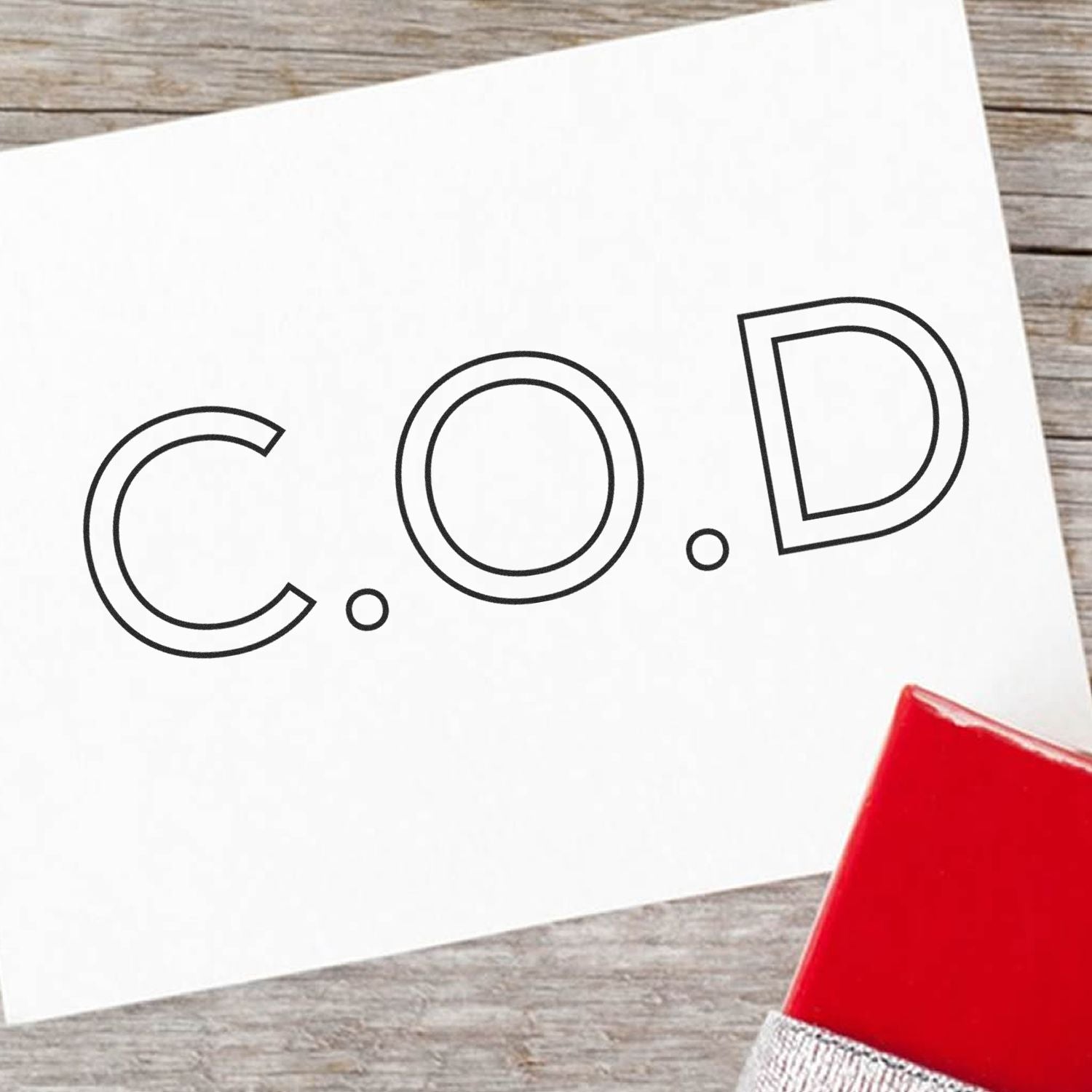 A white card stamped with C.O.D using the Self Inking COD Outline Stamp, placed on a wooden surface with a red object nearby.