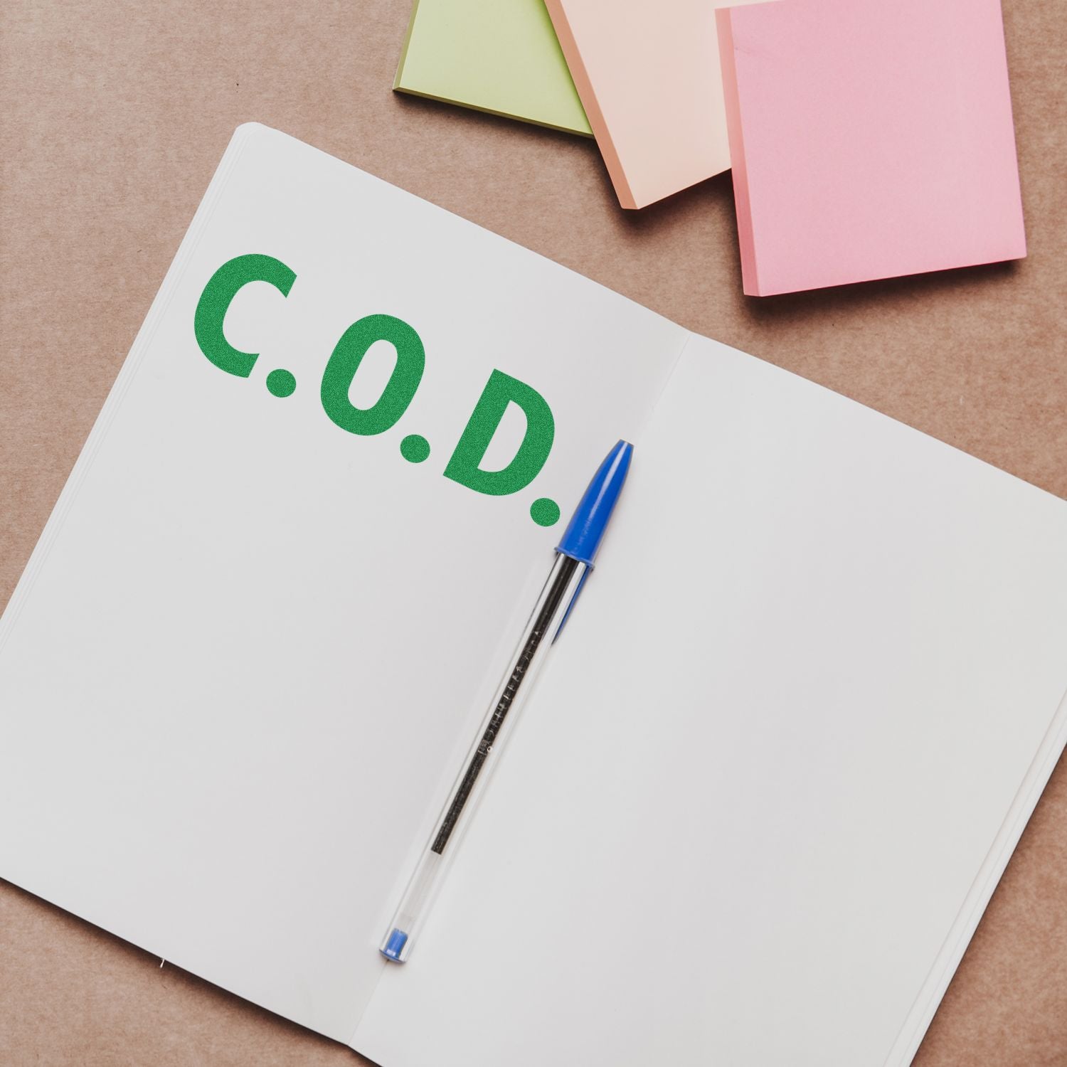 Open notebook stamped with C.O.D. in green using a COD Rubber Stamp, with a blue pen and colorful sticky notes on a brown surface.