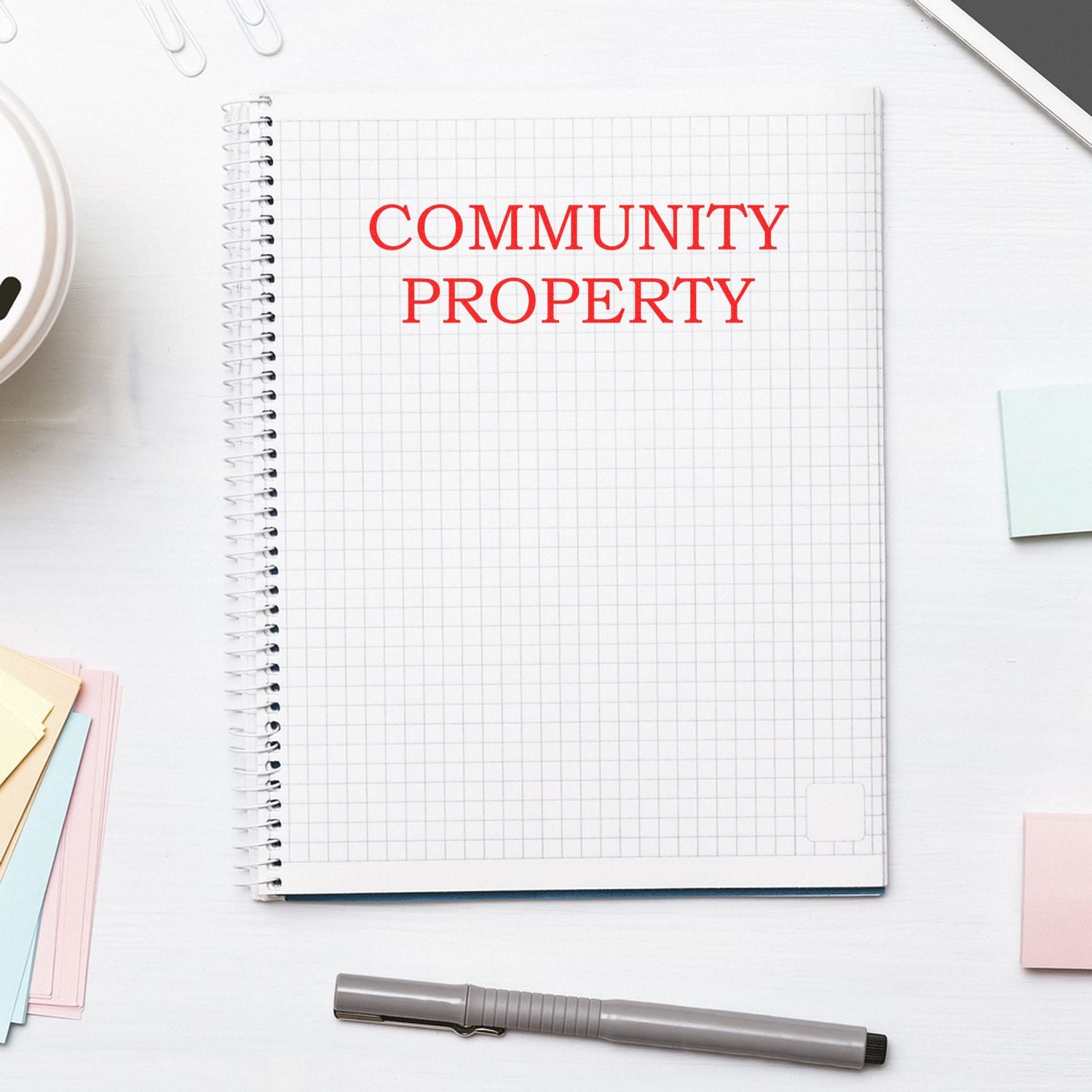 A notebook with Community Property stamped in red using the Community Property Rubber Stamp, surrounded by office supplies on a white desk.