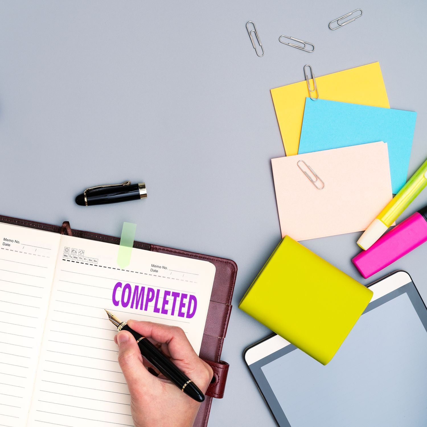 Hand using a Completed Rubber Stamp on a planner, surrounded by colorful sticky notes, paper clips, a pen, highlighters, and a tablet.