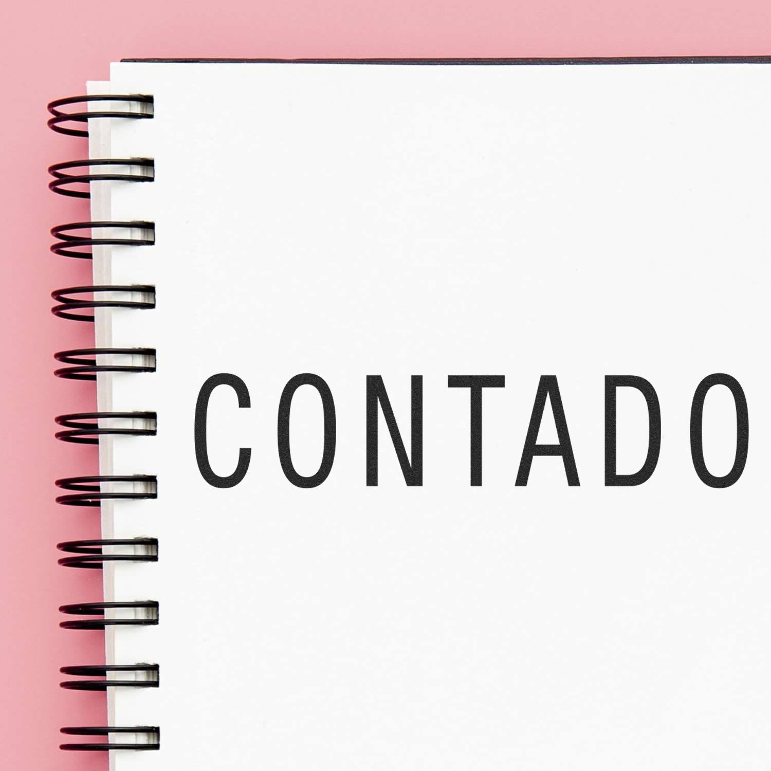 Contado Rubber Stamp imprint on a white spiral notebook with a pink background.