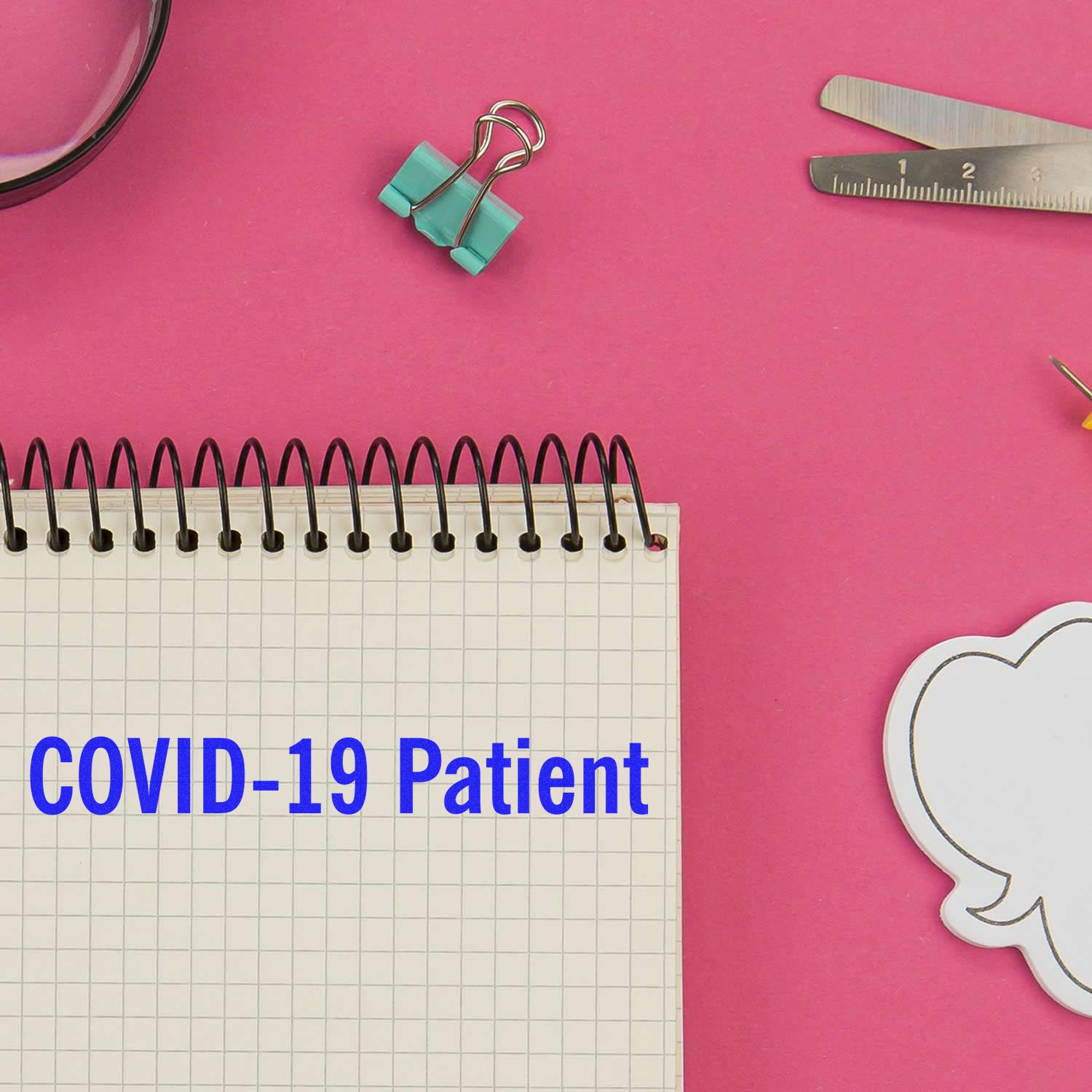 A rubber stamp with COVID-19 Patient text on a spiral notebook, surrounded by office supplies on a pink background.