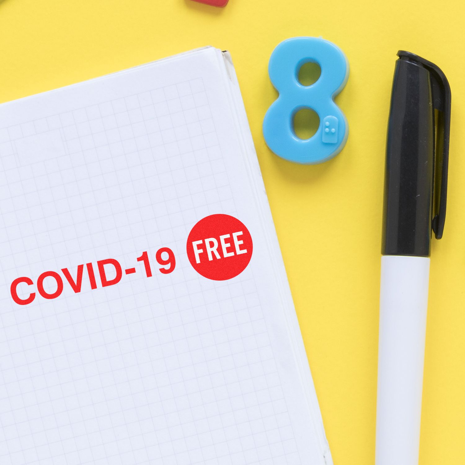 Covid-19 Rubber Stamp marking COVID-19 FREE in red on a notebook, with a pen and a blue number 8 magnet on a yellow background.