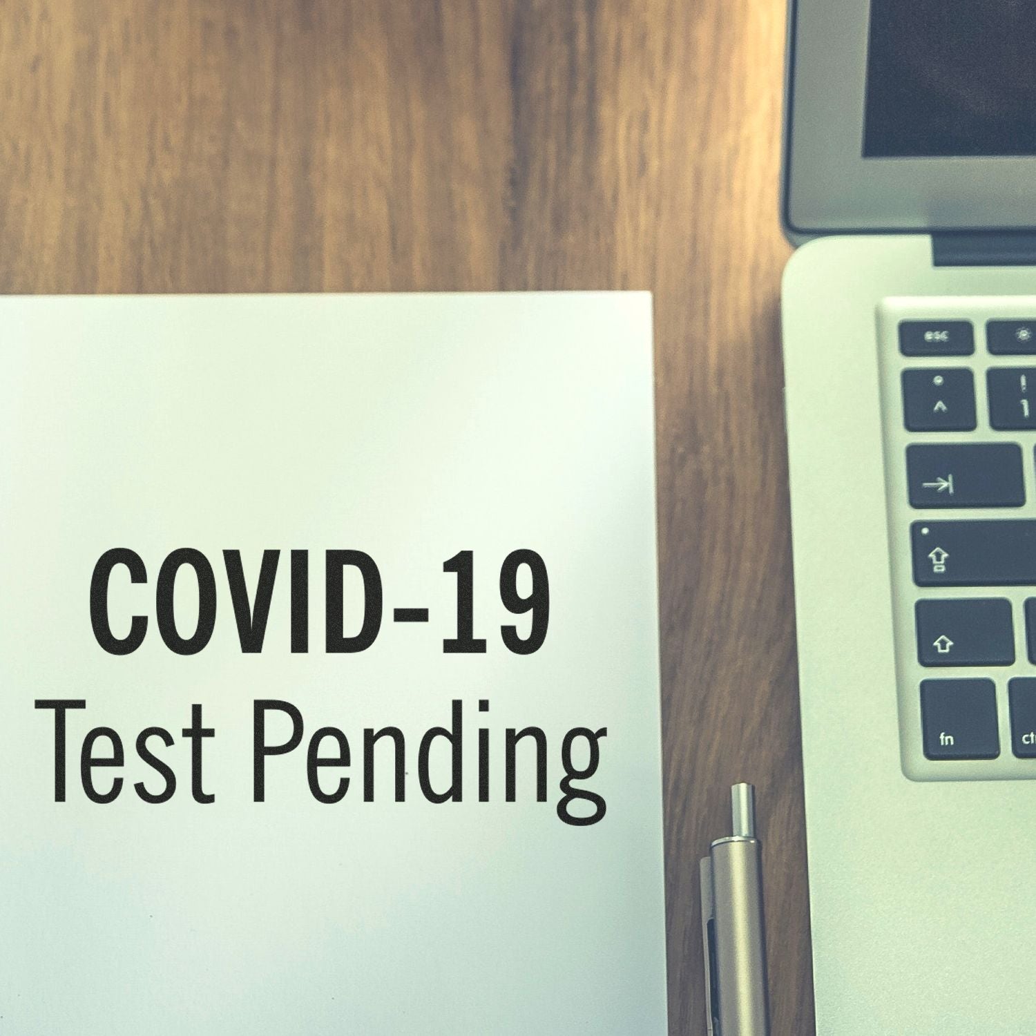 A paper stamped with COVID-19 Test Pending using the Large Self Inking Covid-19 Test Pending Stamp, next to a laptop and pen on a wooden desk.