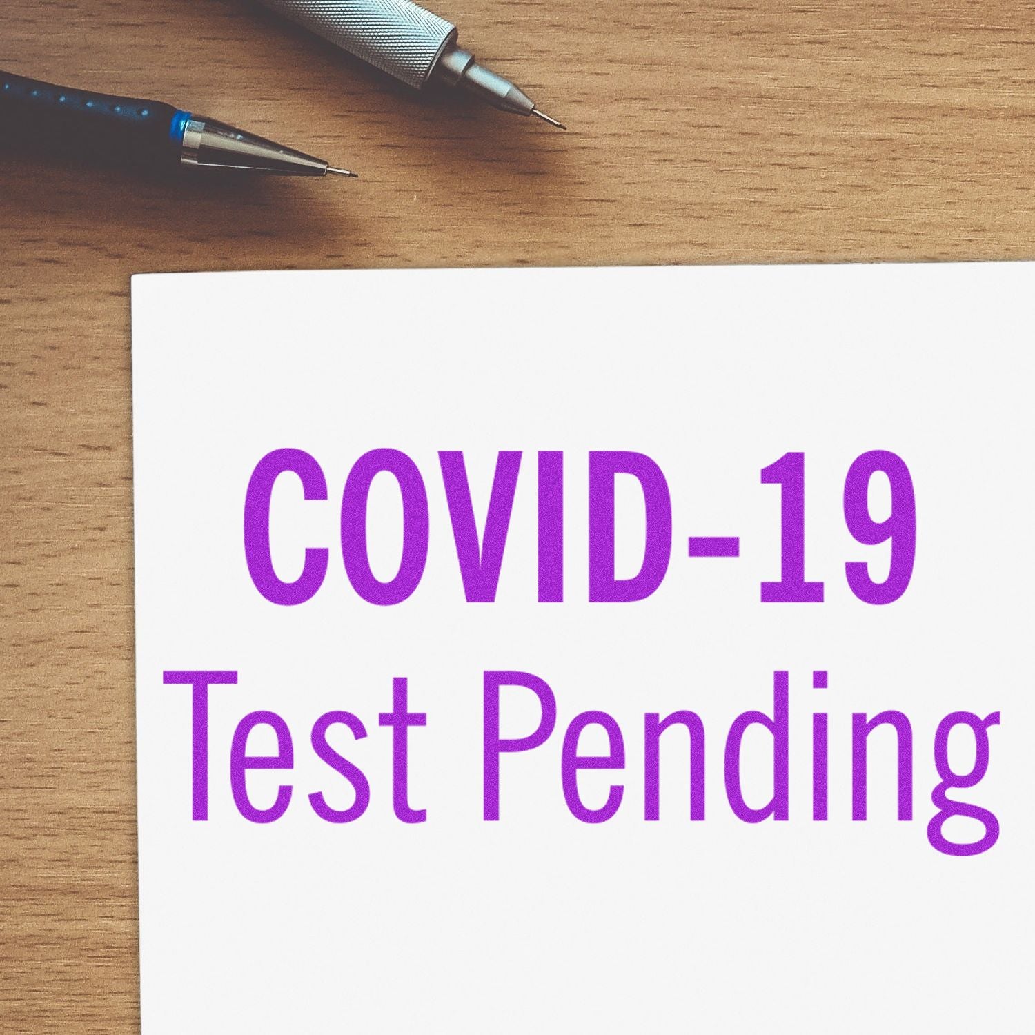 Self Inking Covid-19 Test Pending Stamp in use, showing the stamped text COVID-19 Test Pending on paper, with pens on a wooden desk.