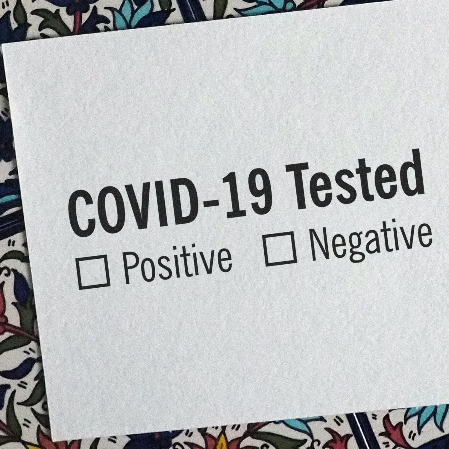 Covid-19 Tested rubber stamp with checkboxes for Positive and Negative on a white paper, placed on a floral background.