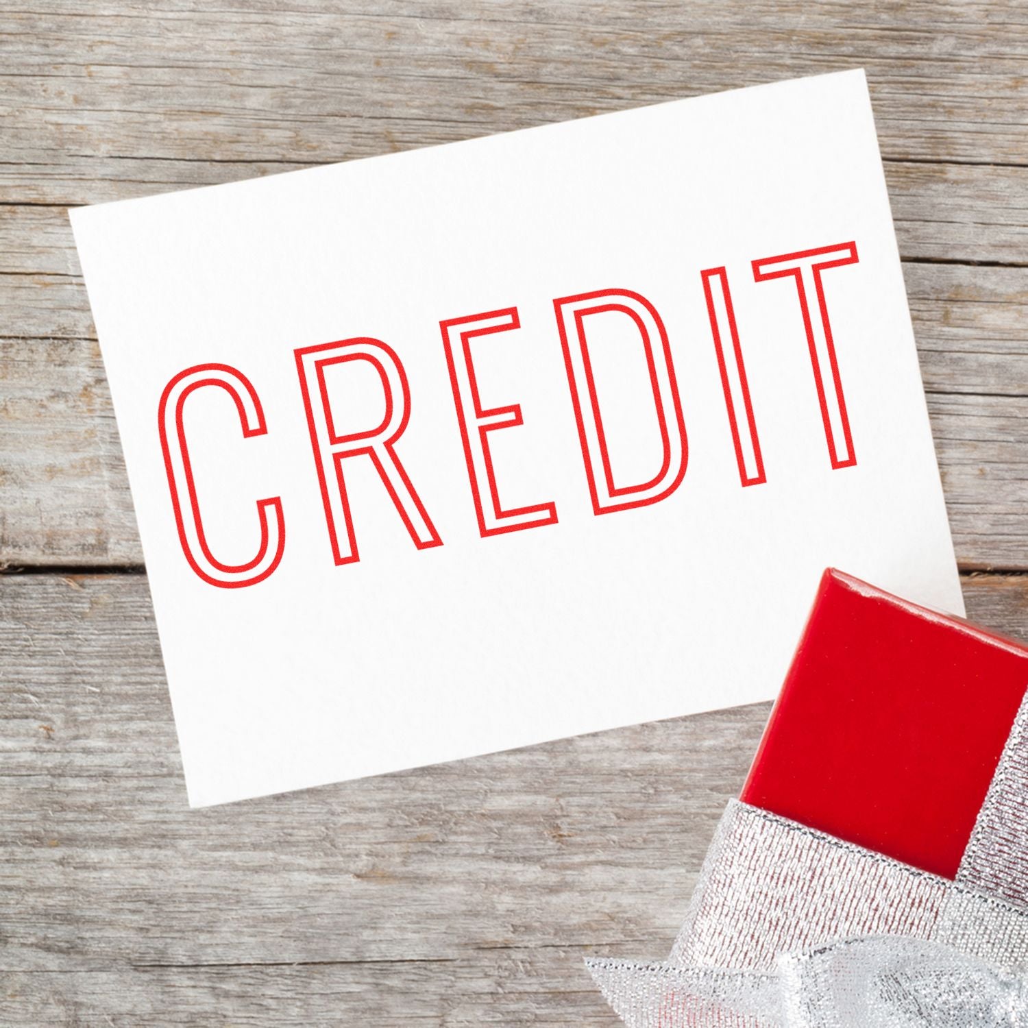 A white card stamped with CREDIT in red using the Credit Outline Rubber Stamp, placed on a wooden surface next to a red gift box.