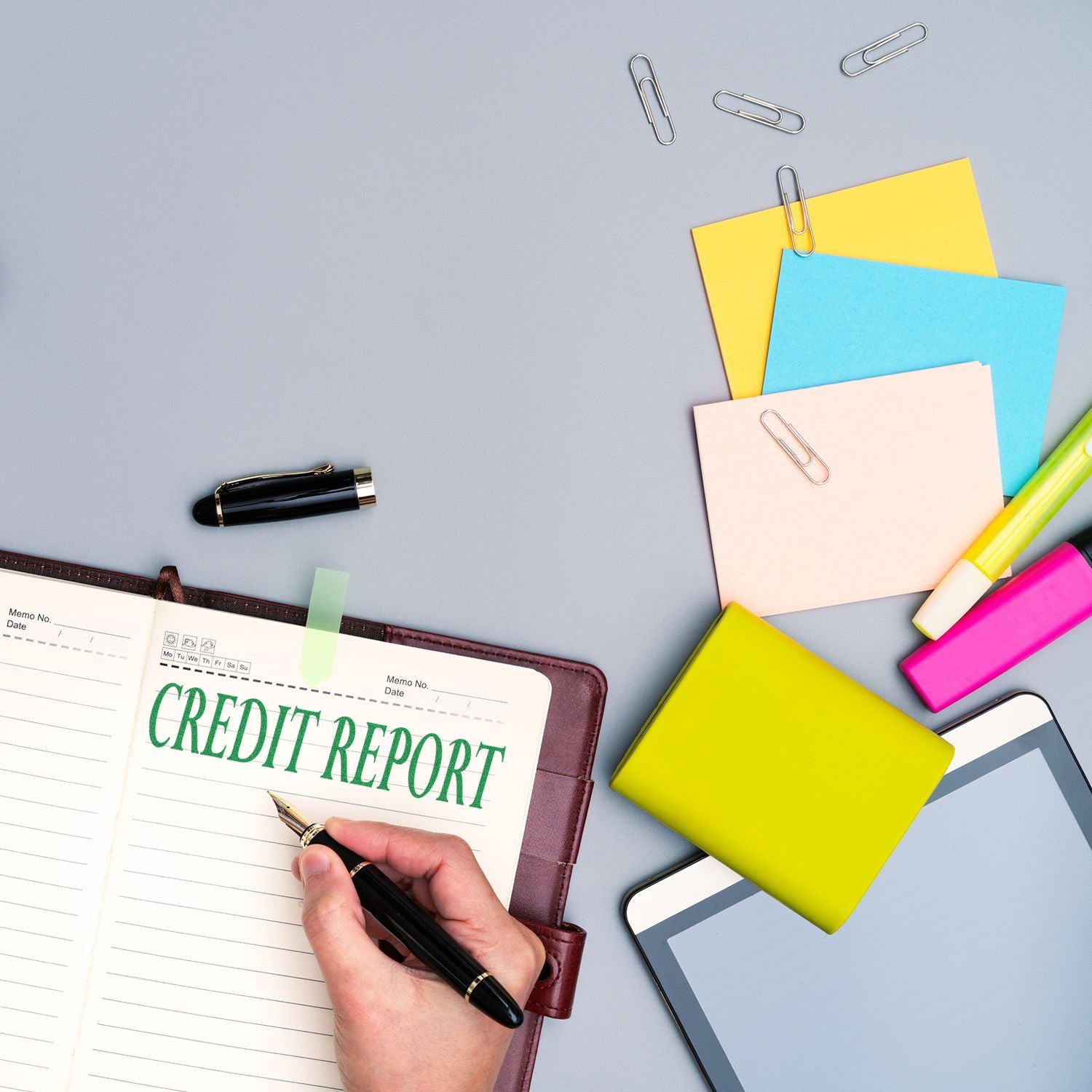 Hand using a Credit Report Rubber Stamp on a notebook, surrounded by colorful sticky notes, paper clips, a pen, and a tablet.