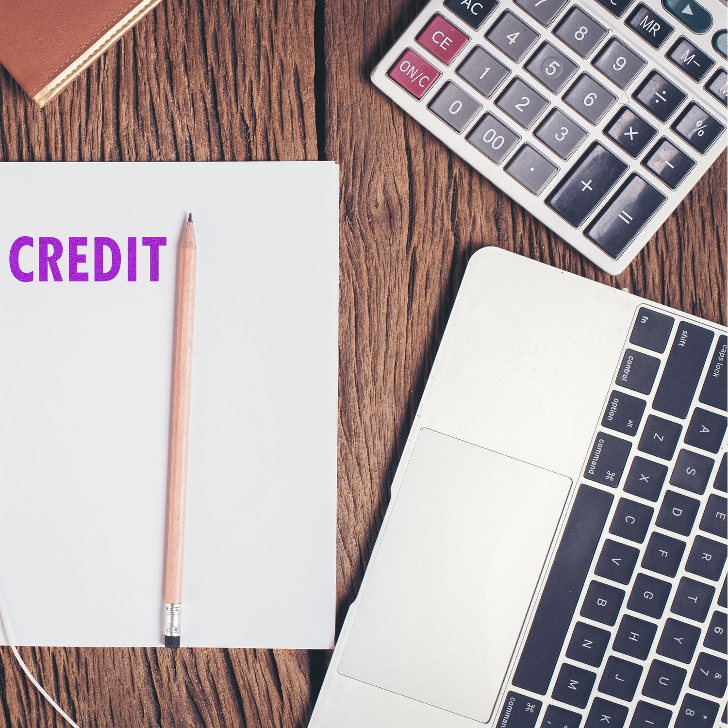 A Large Self Inking Credit Stamp marks CREDIT on a white paper beside a pencil, calculator, and laptop on a wooden desk.