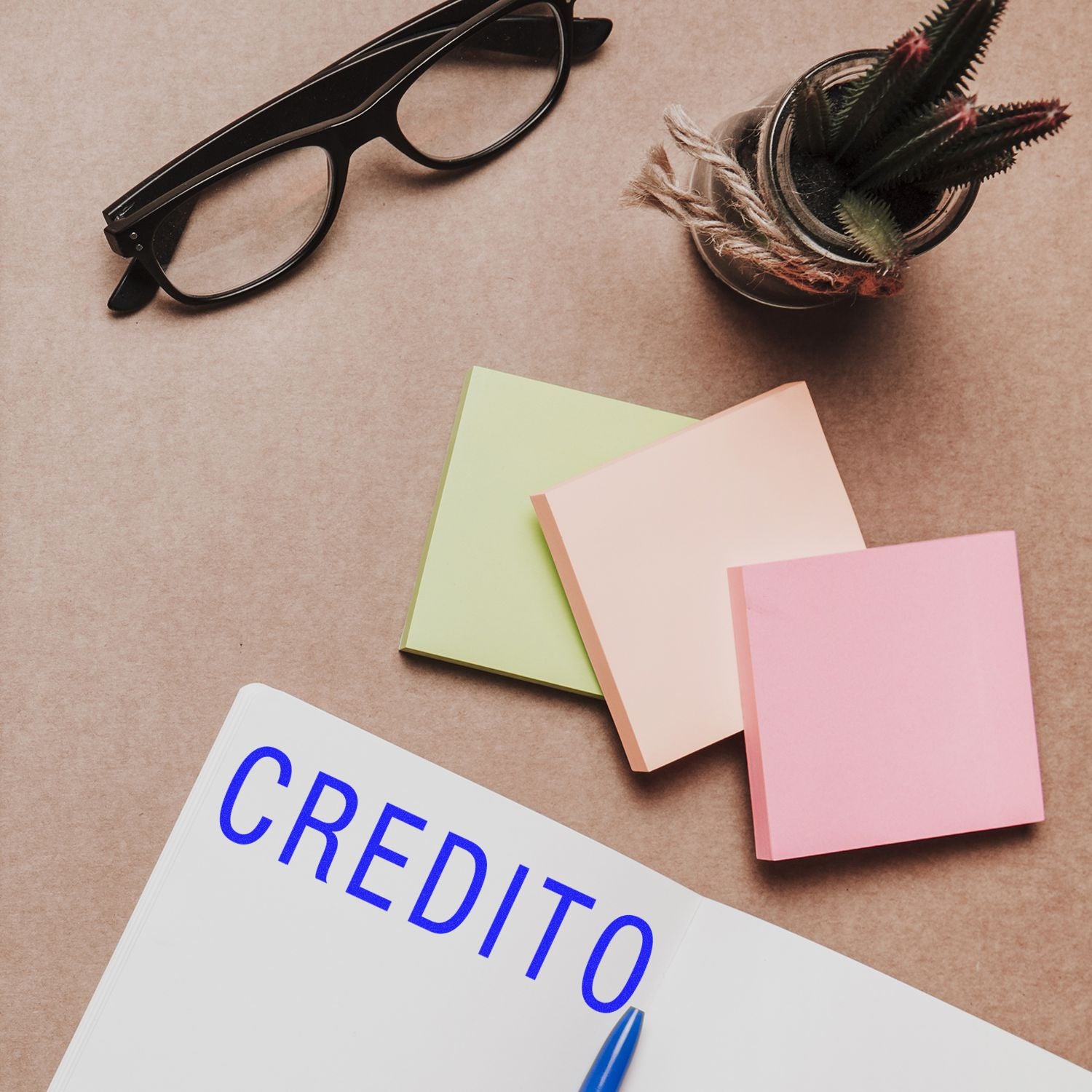 Large Credito Rubber Stamp in use on a notebook, surrounded by sticky notes, a pen, glasses, and a small potted plant on a desk.