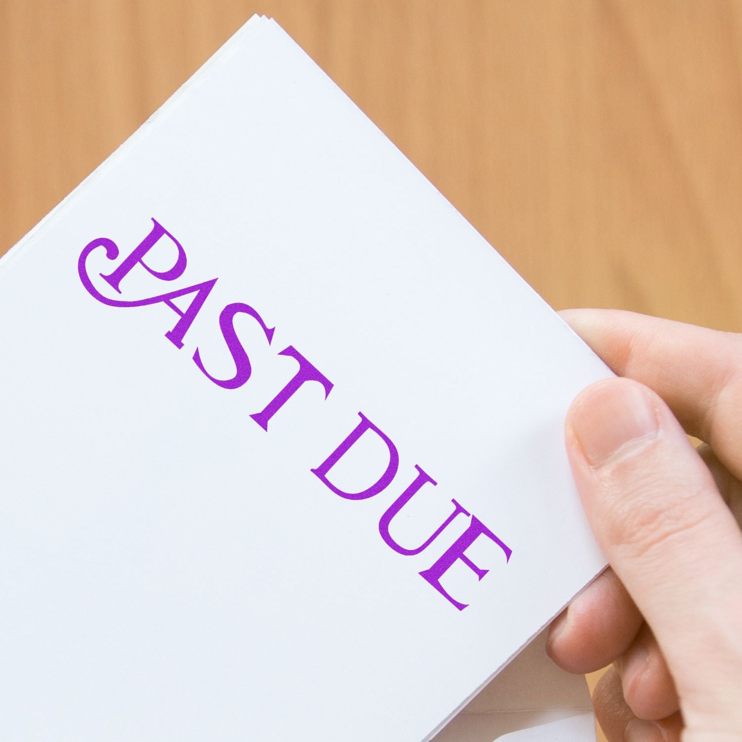 Hand holding a paper stamped with PAST DUE in purple ink using the Curley Past Due Rubber Stamp, against a wooden background.