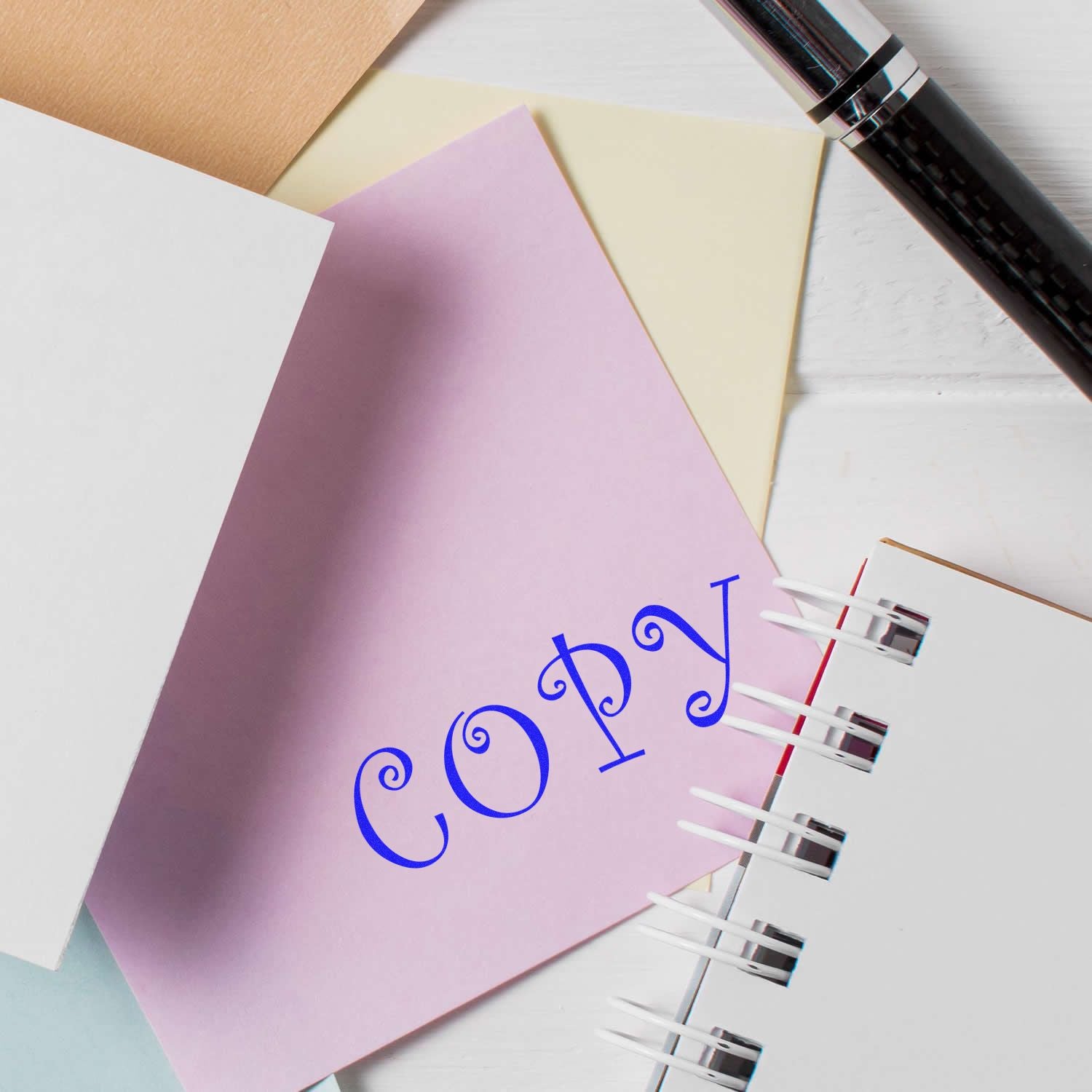 Curly Copy Rubber Stamp imprint on pastel paper, surrounded by stationery items including a pen, notebook, and additional sheets of paper.