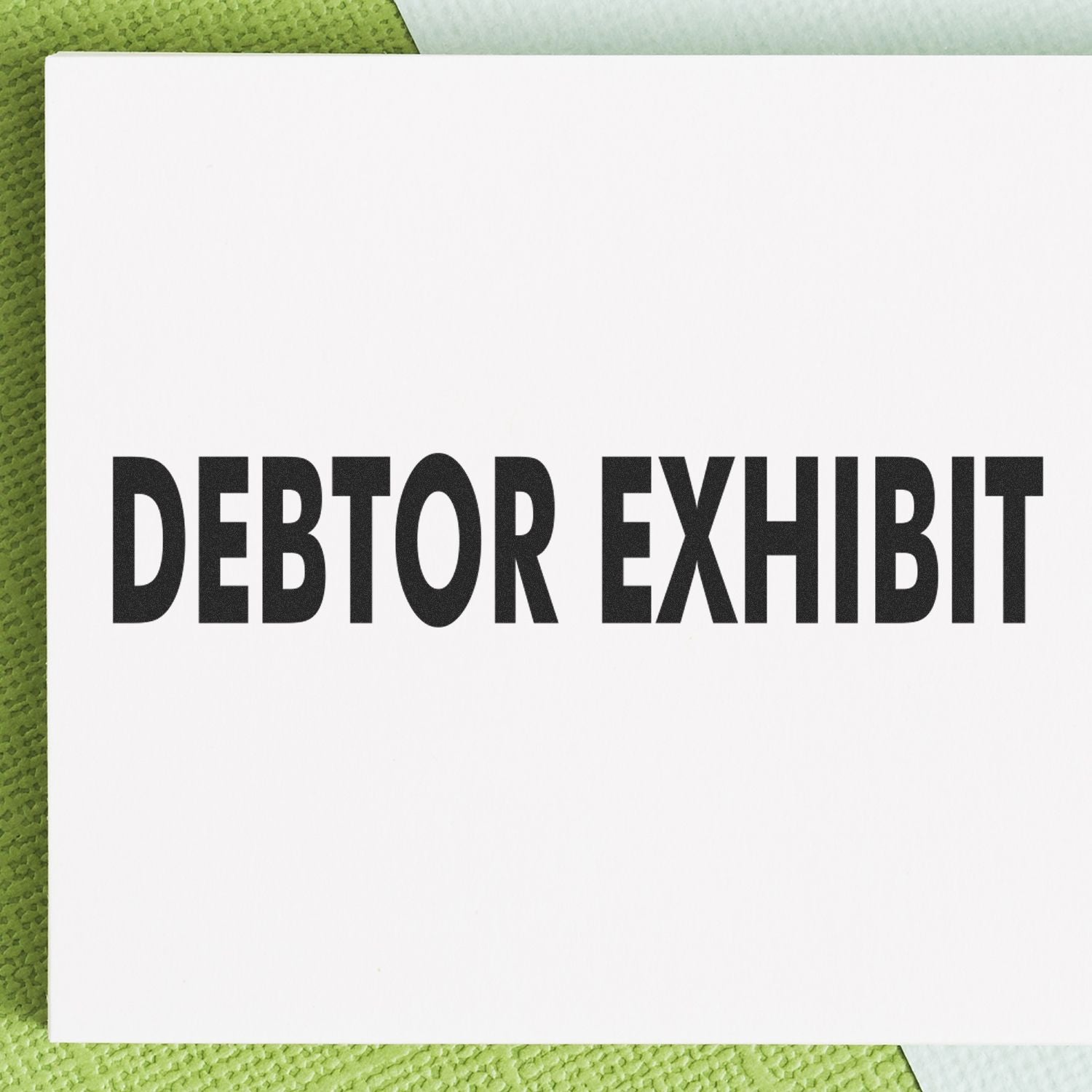 Debtor Exhibit Rubber Stamp in use, displaying bold black text DEBTOR EXHIBIT on a white background with a green textured surface underneath.