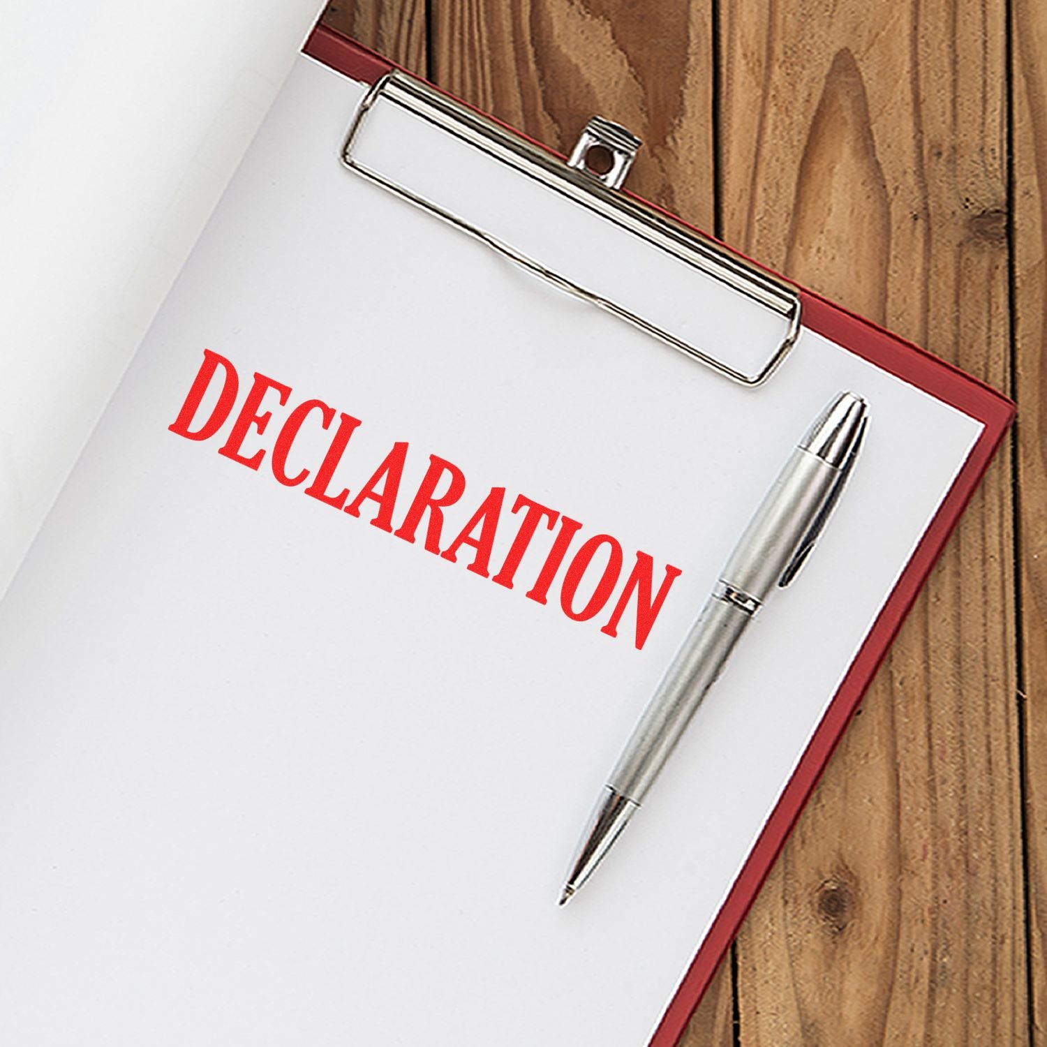 Self Inking Declaration Stamp imprinting DECLARATION in red on a white paper on a clipboard with a silver pen, placed on a wooden surface.