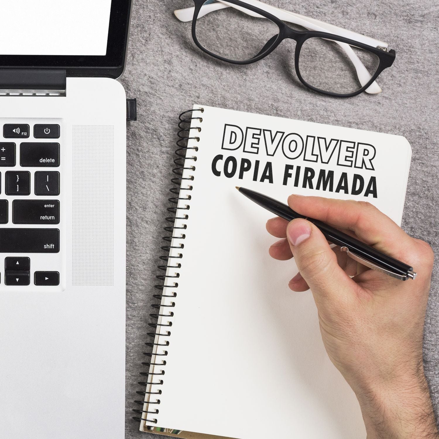 Hand holding a pen next to a notebook stamped with DEVOLVER COPIA FIRMADA using the Devolver Copia Rubber Stamp, beside a laptop and glasses.