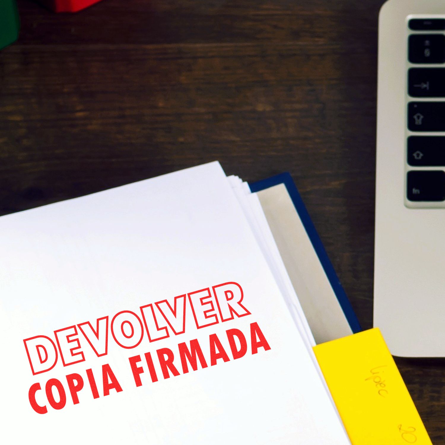 A stack of papers stamped with DEVOLVER COPIA FIRMADA using the Self Inking Devolver Copia Stamp, next to a laptop on a wooden desk.