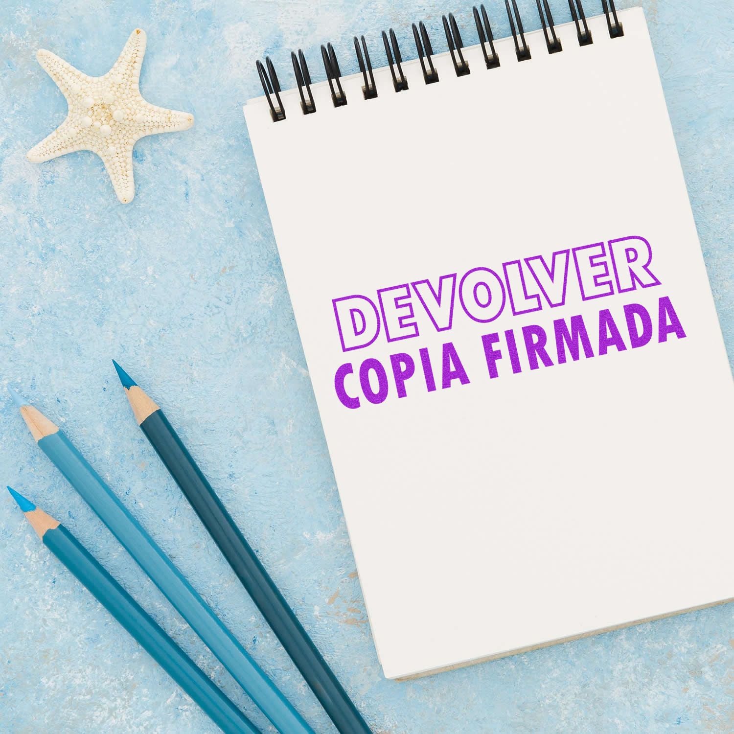 Devolver Copia Rubber Stamp used on a spiral notebook with 'DEVOLVER COPIA FIRMADA' text, surrounded by blue pencils and a starfish.