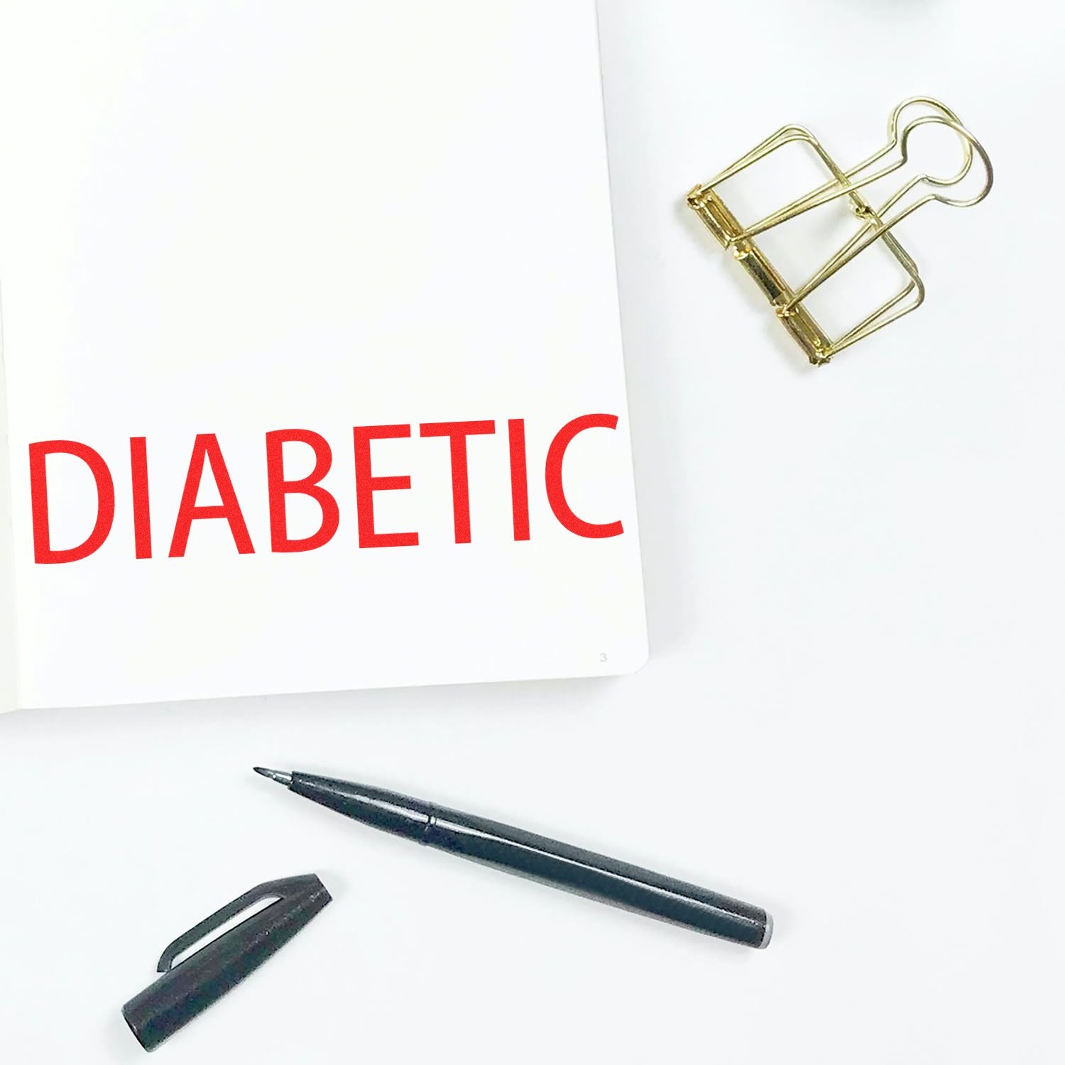 Large Pre-Inked Diabetic Stamp in use, marking DIABETIC in red on a white page, with a pen, cap, and gold binder clip nearby.