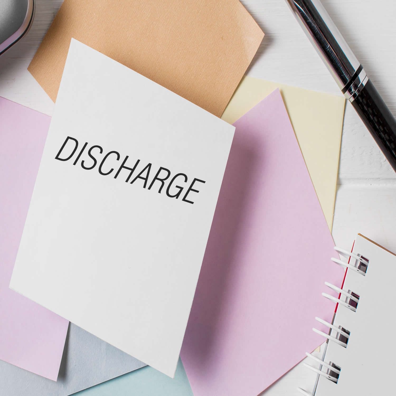 Discharge Rubber Stamp in use on a white paper surrounded by pastel-colored sheets, a pen, and a spiral notebook on a white surface.