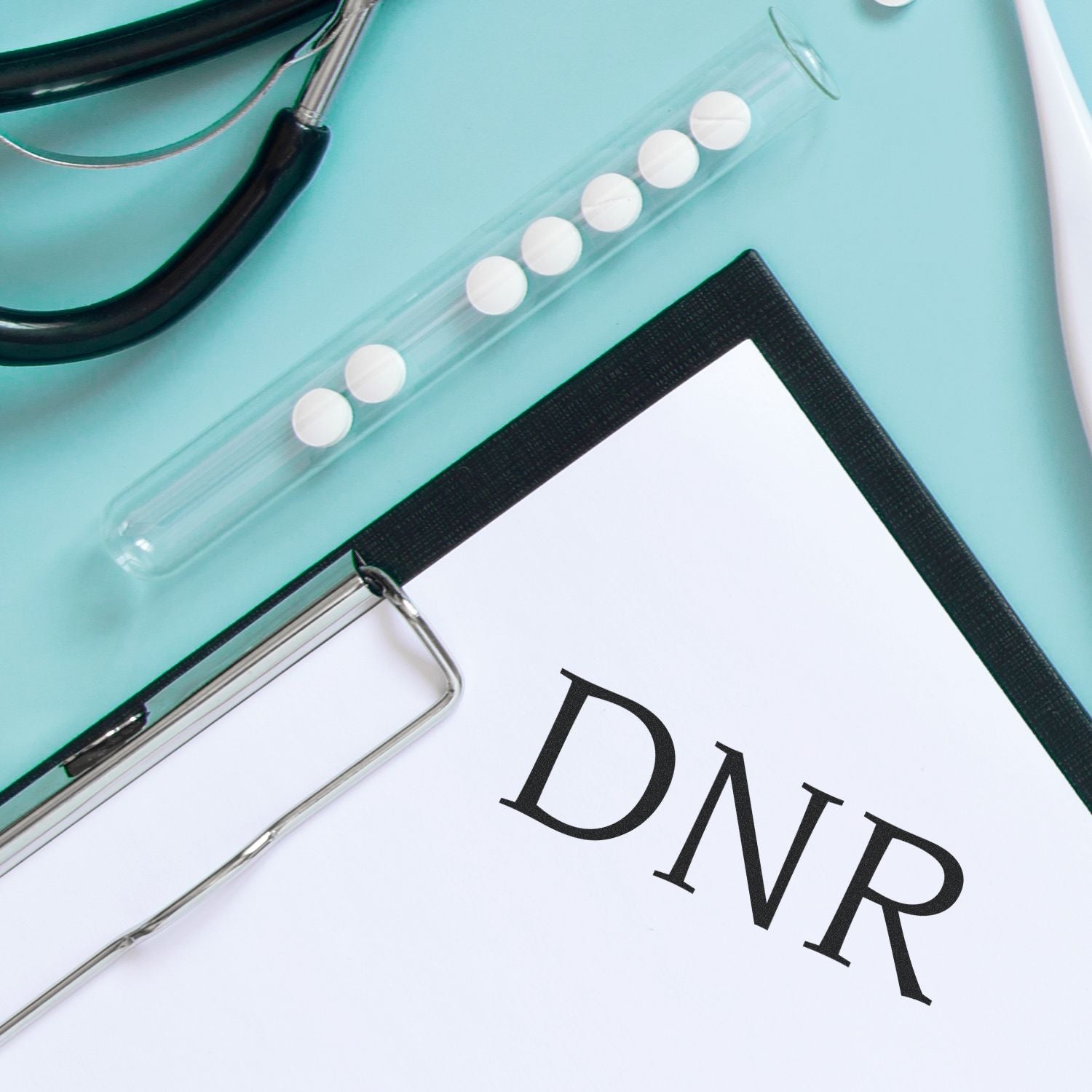 A DNR Medical Rubber Stamp next to a stethoscope, test tube with pills, and a clipboard with a DNR form on a light blue surface.