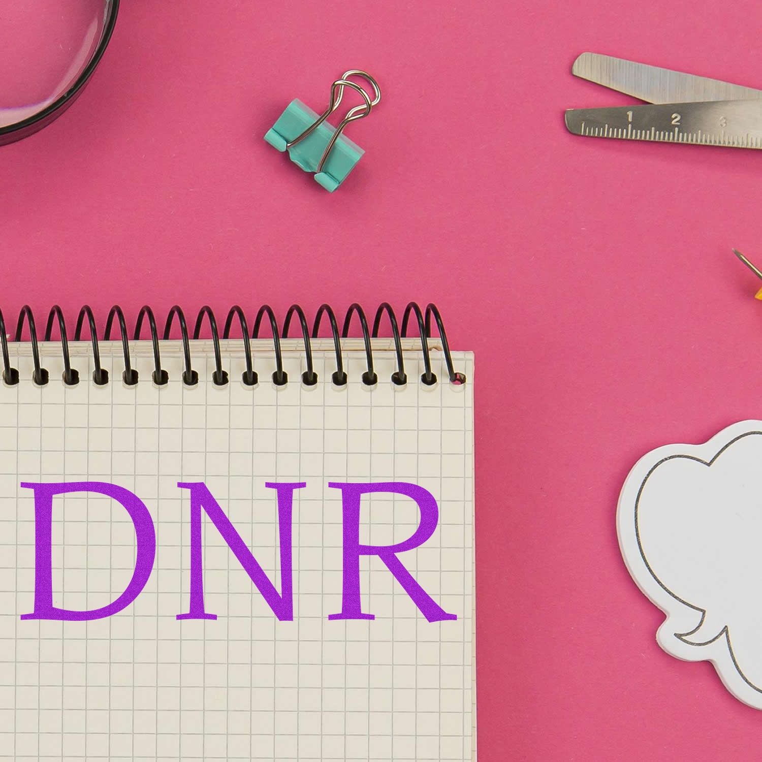 A DNR Medical Rubber Stamp is shown in use on a spiral notebook with a pink background, surrounded by office supplies.