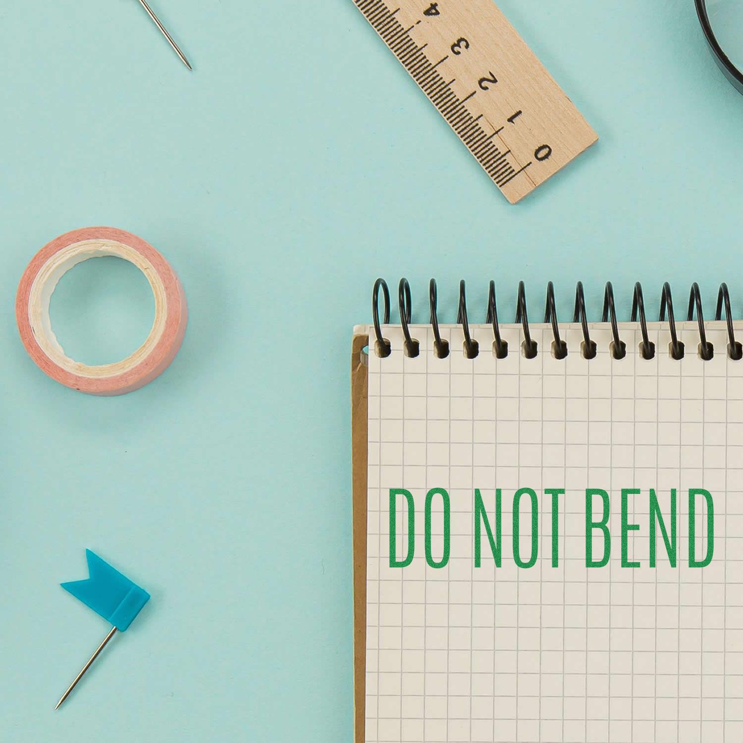 Large Self Inking Do Not Bend Stamp used on a spiral notebook, surrounded by a ruler, tape, and a blue flag pin on a light blue background.