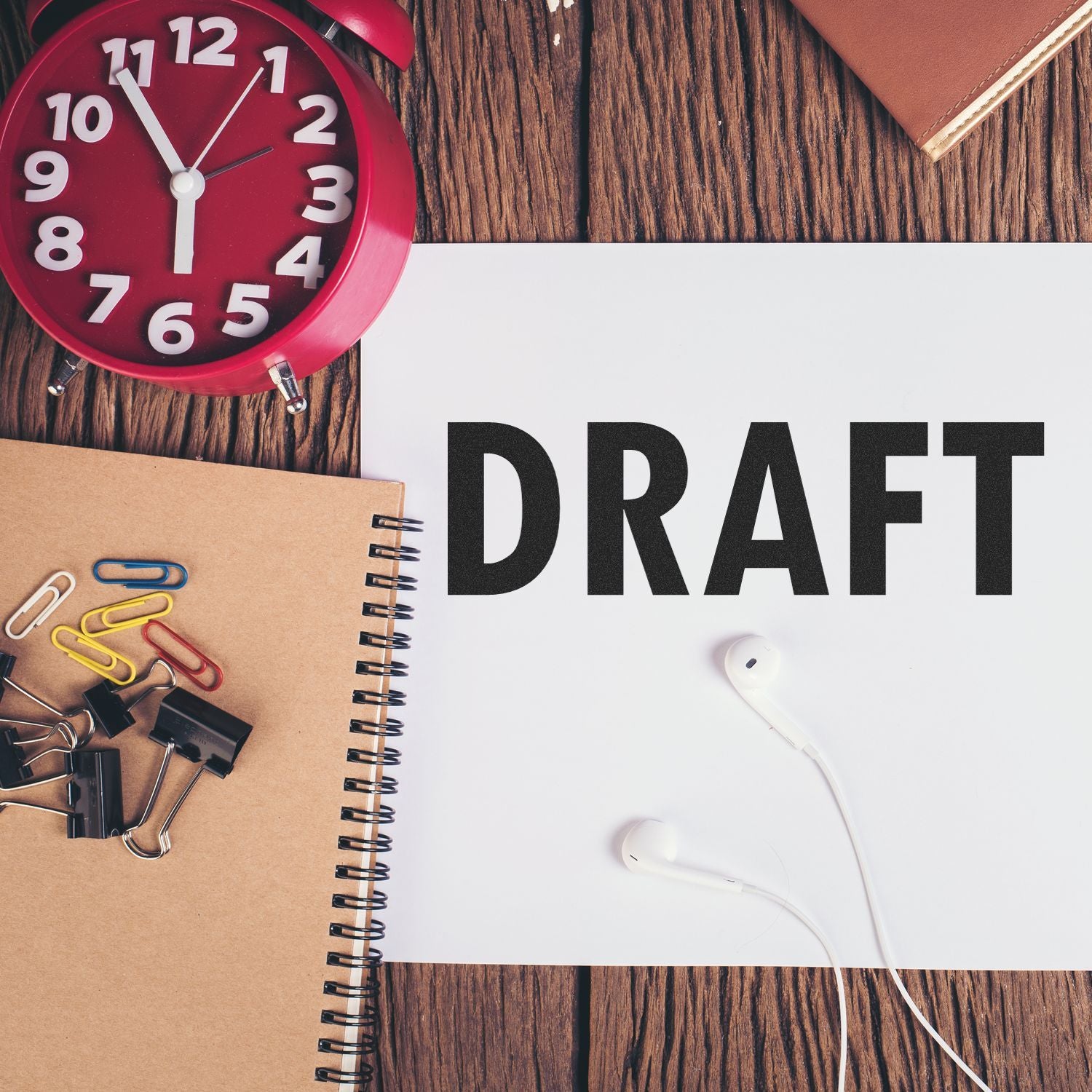 A desk with a clock, notebook, paper clips, and earbuds, featuring a paper stamped with DRAFT using the Draft Rubber Stamp.