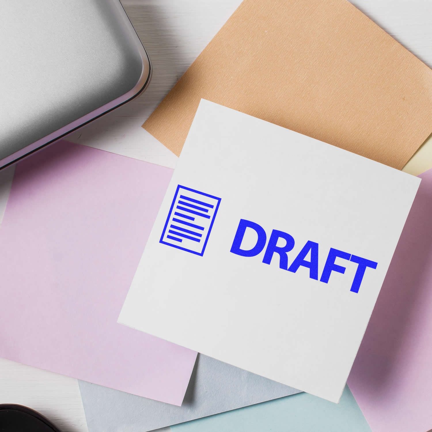 Draft with Letter Rubber Stamp in use on a white paper, surrounded by pastel-colored sheets and a silver laptop on a white desk.